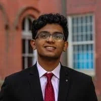 A young man in business attire with glasses, smiling outdoors in front of a building with red and blue windows.