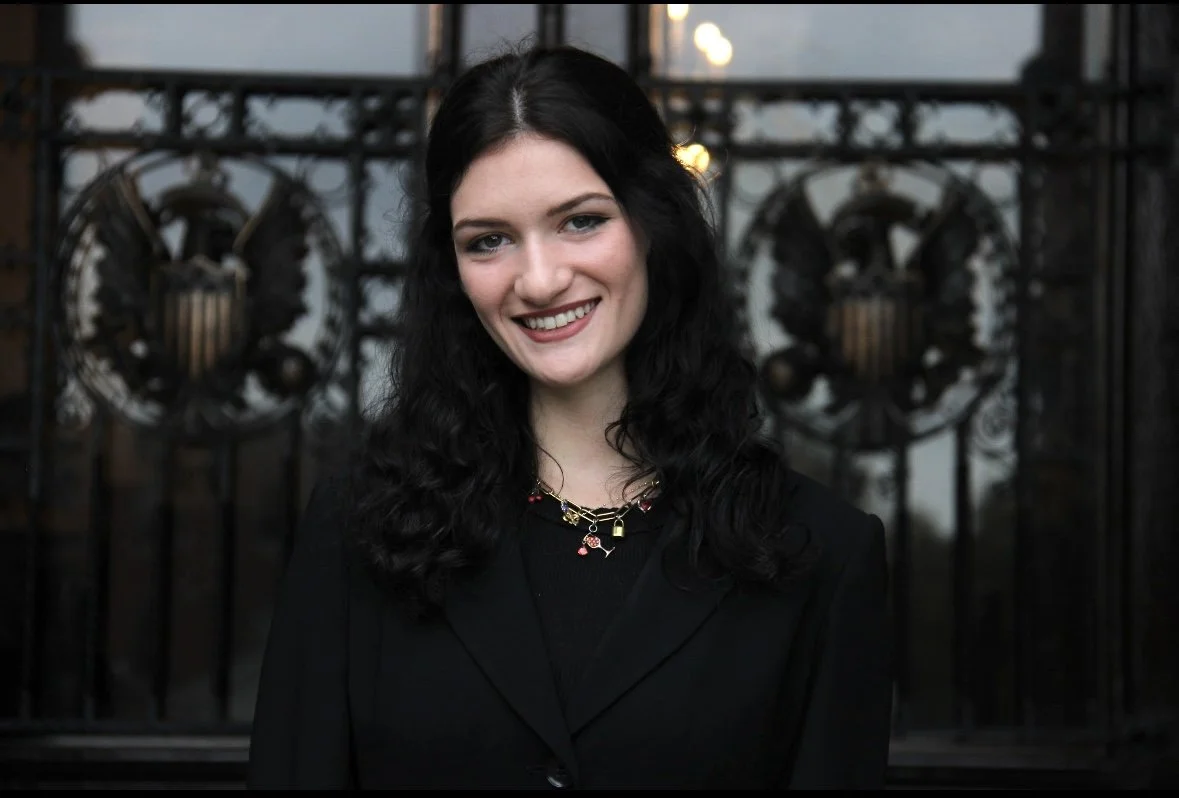 Young woman with dark wavy hair smiling, wearing a black blazer and a gold necklace, standing outdoors in front of an ornate wrought iron gate.