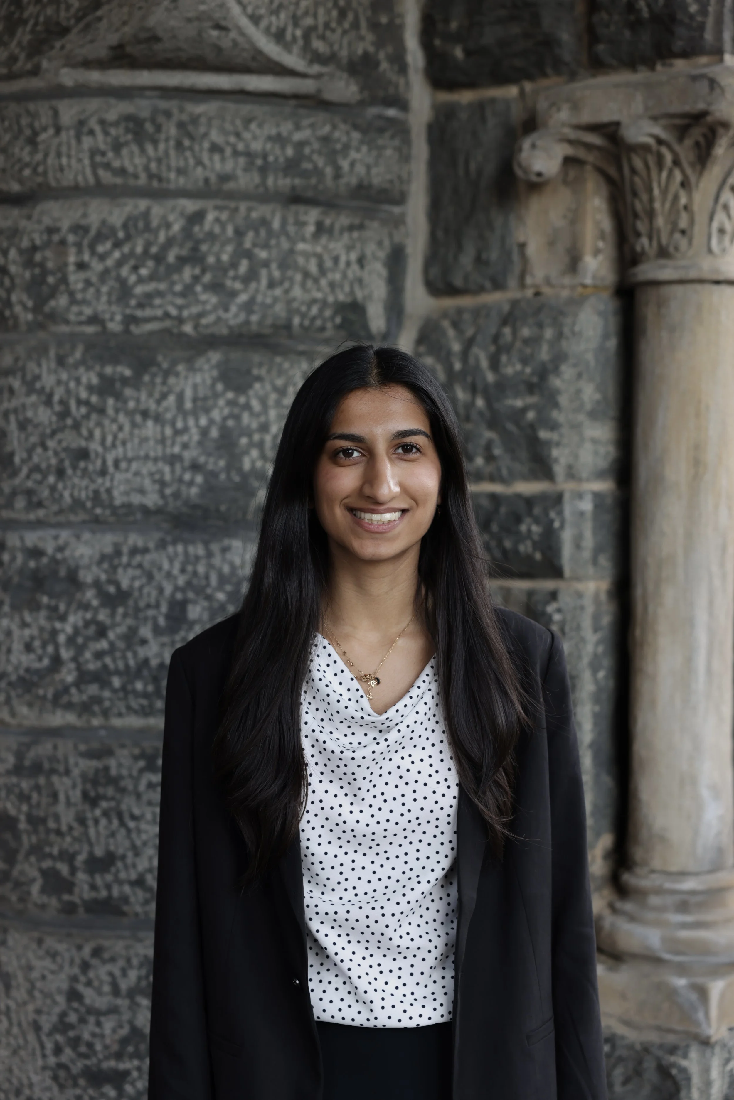 A woman with long dark hair smiling, wearing a black blazer and a white blouse with black polka dots, standing in front of a historic stone building with carved columns.