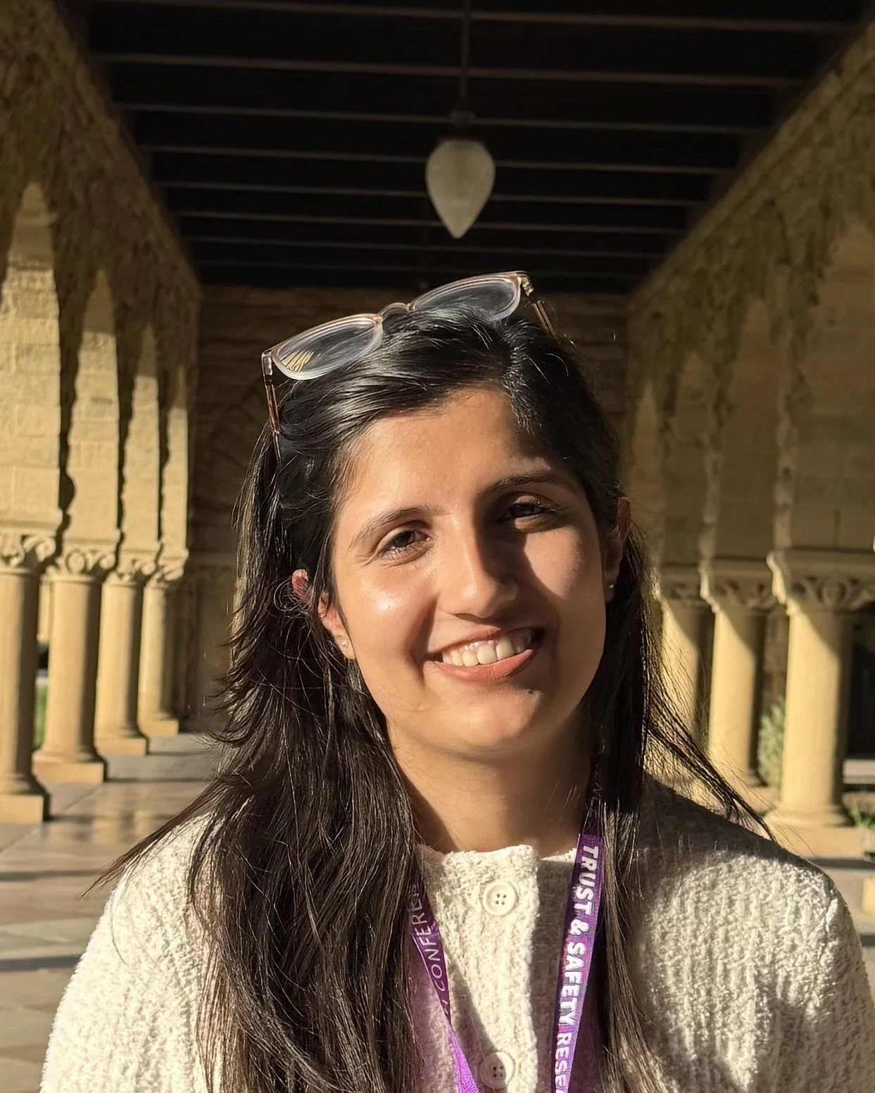 Young woman smiling with glasses on her head, standing in a sunny outdoor corridor with columns and arches.