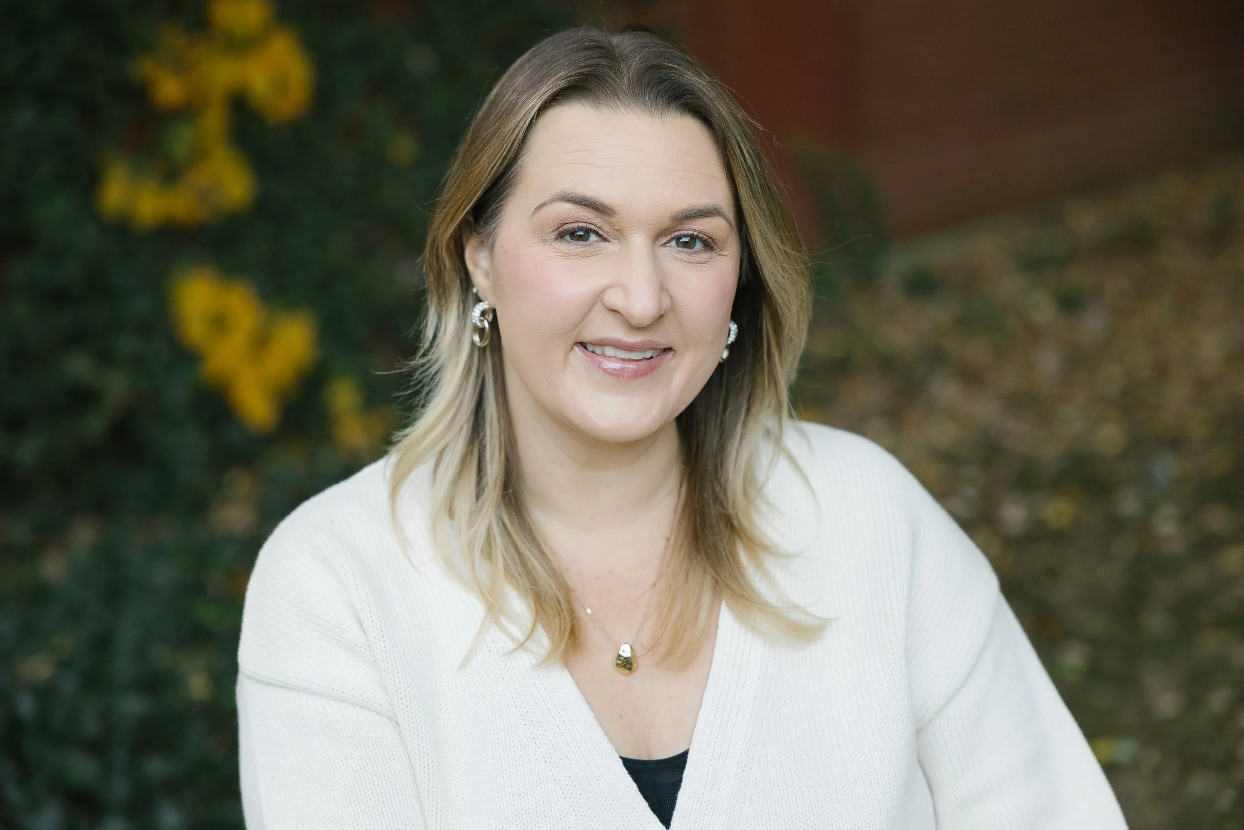 A smiling woman with shoulder-length blonde hair, wearing earrings, a necklace, and a white cardigan, outdoors with greenery and yellow flowers in the background.