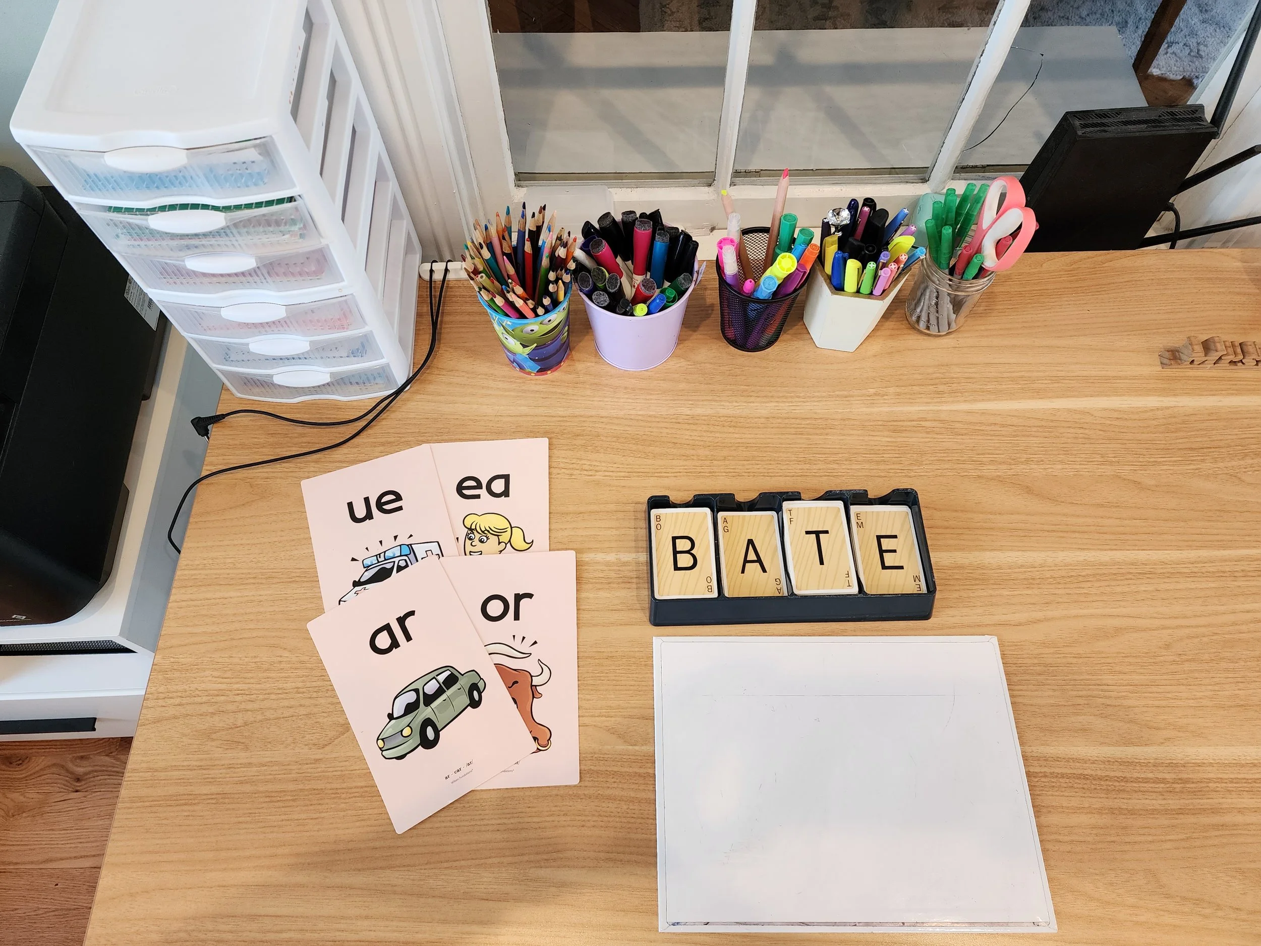 Desk with various coloring supplies, flashcards with pictures of a police car and a dog with the words 'ar' and 'or', and letter tiles spelling 'BATE'.