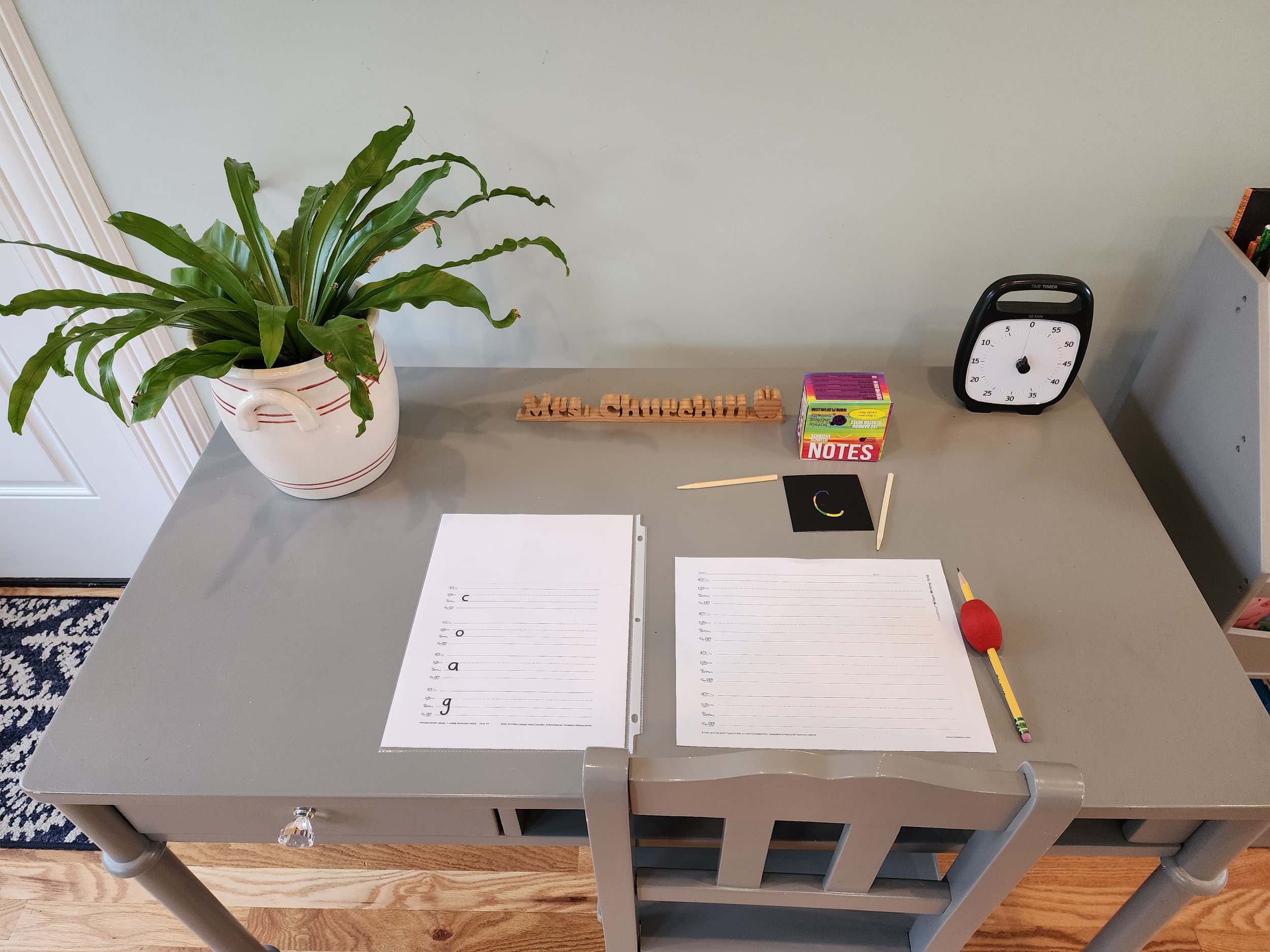 A desk with a potted plant on the left, two sheets of lined paper labeled 'vocab' and 'spelling' in the center, and various school supplies including a timer, a pack of notes, a small black card, a red and yellow pencil with a balloon-shaped eraser, 