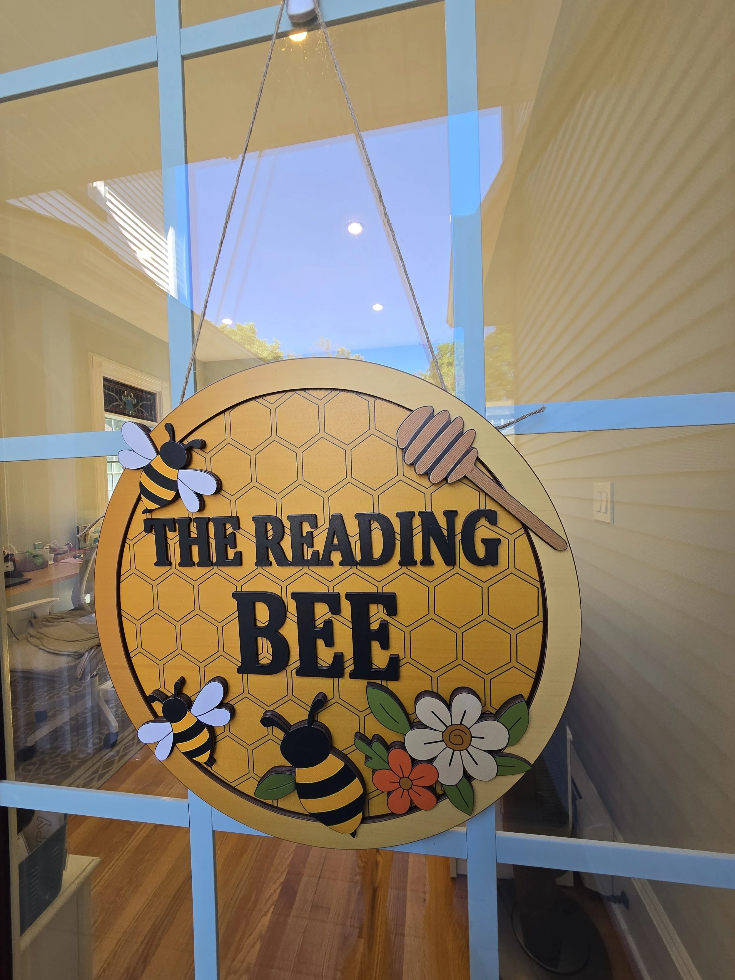 Decorative round sign on glass window with honeycomb pattern, featuring bees, a honey dipper, and flowers, with the text "The Reading Bee".