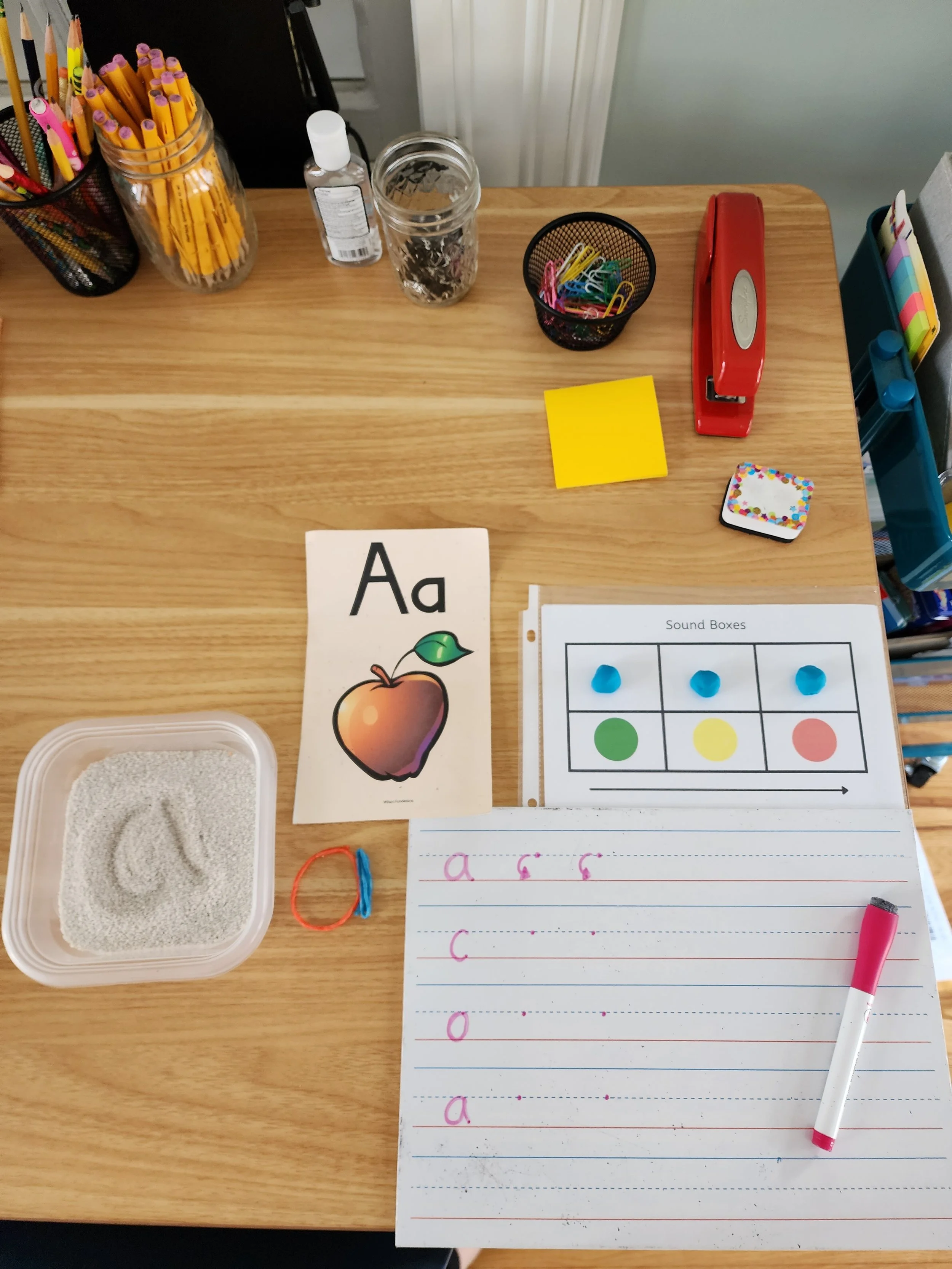Educational desk setup for young learners with alphabet flashcard, sound boxes chart, writing practice, and various stationery items.