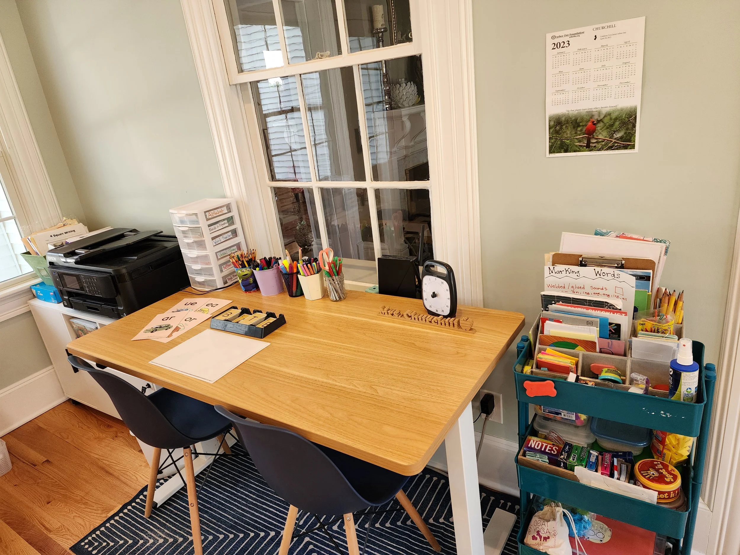A workspace with a wooden table holding containers with colored pencils and markers, a clock, and papers. To the left, a printer with paper on top. To the right, a rolling cart filled with school supplies and books. Behind, a window and a calendar on