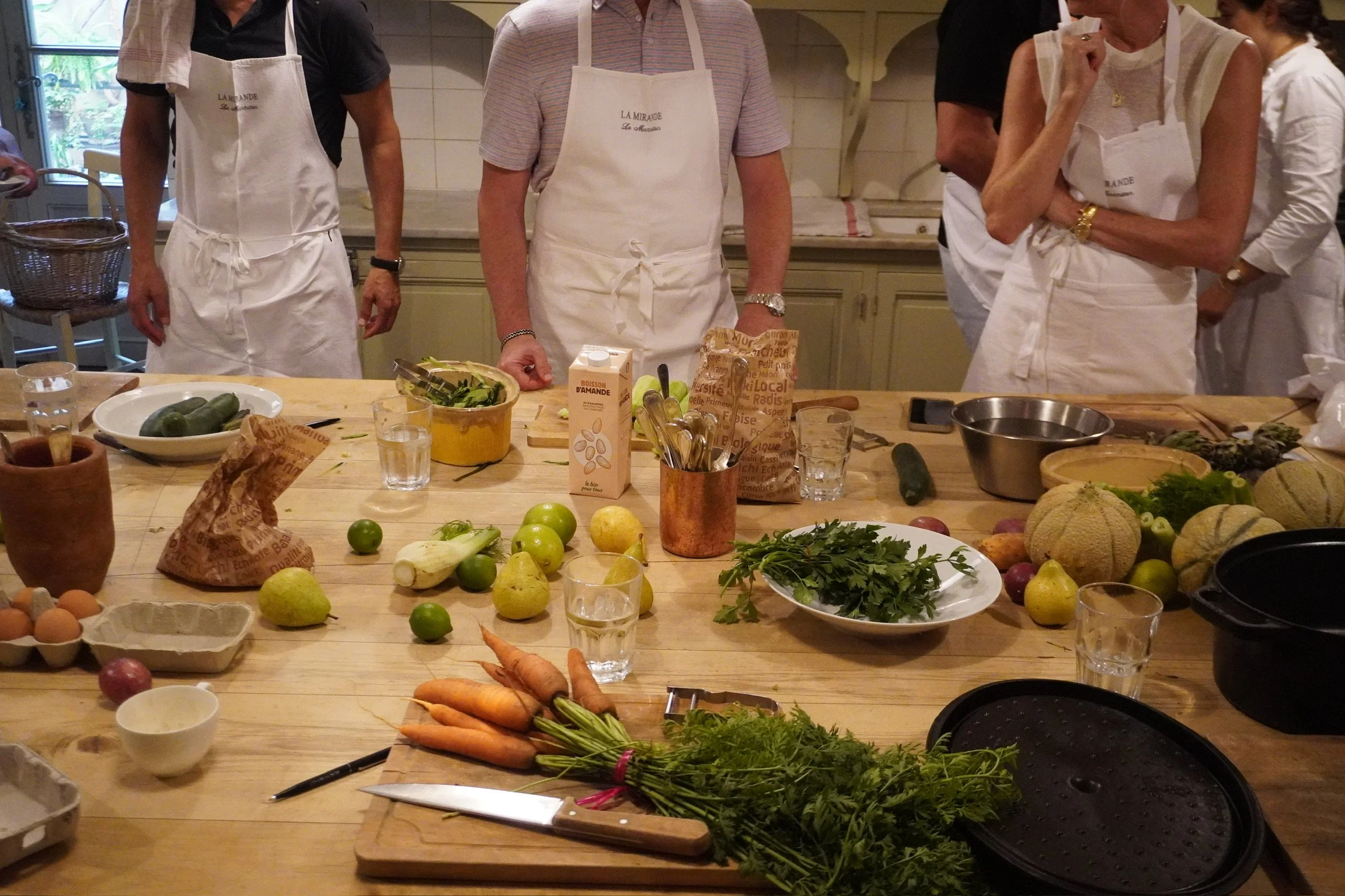 Groupe de personnes portant des tabliers blancs autour d'une table en cuisine, avec des légumes, des fruits, des boîtes, des verres d'eau, et divers ustensiles de cuisine.