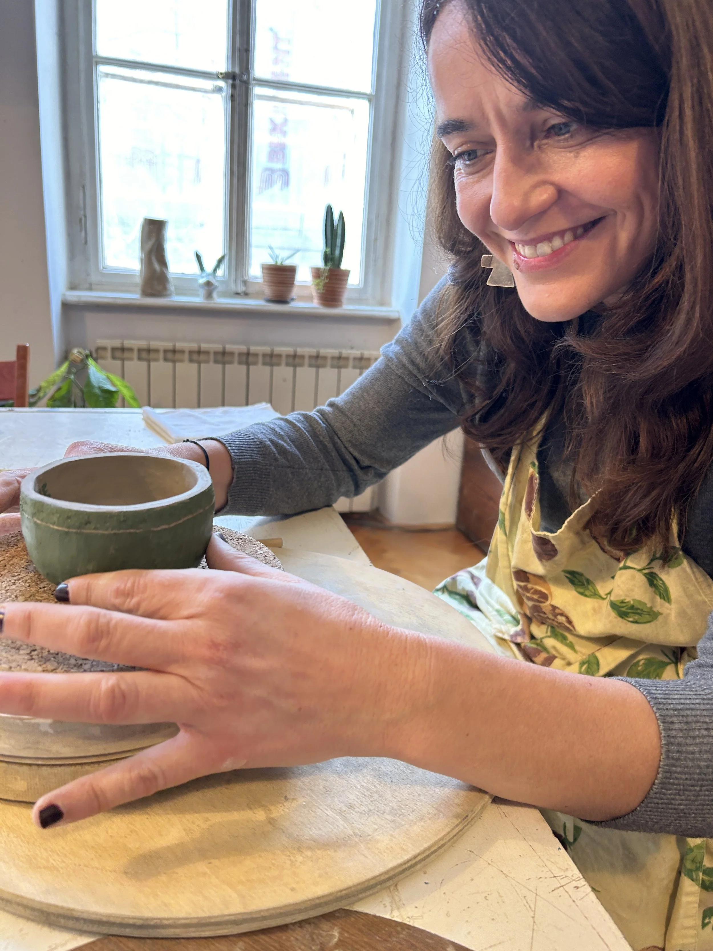 A woman is smiling and working on a ceramic pot in a cozy, well-lit room with a window, potted plants on the windowsill, and a wooden table.