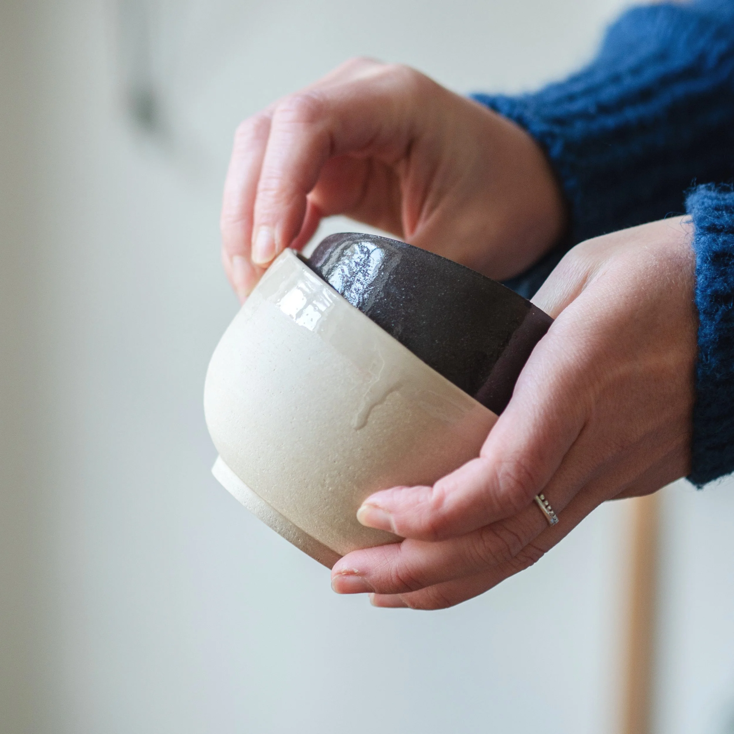 Close-up of a person's hand with a ring holding a ceramic cup or mug, against a plain background.