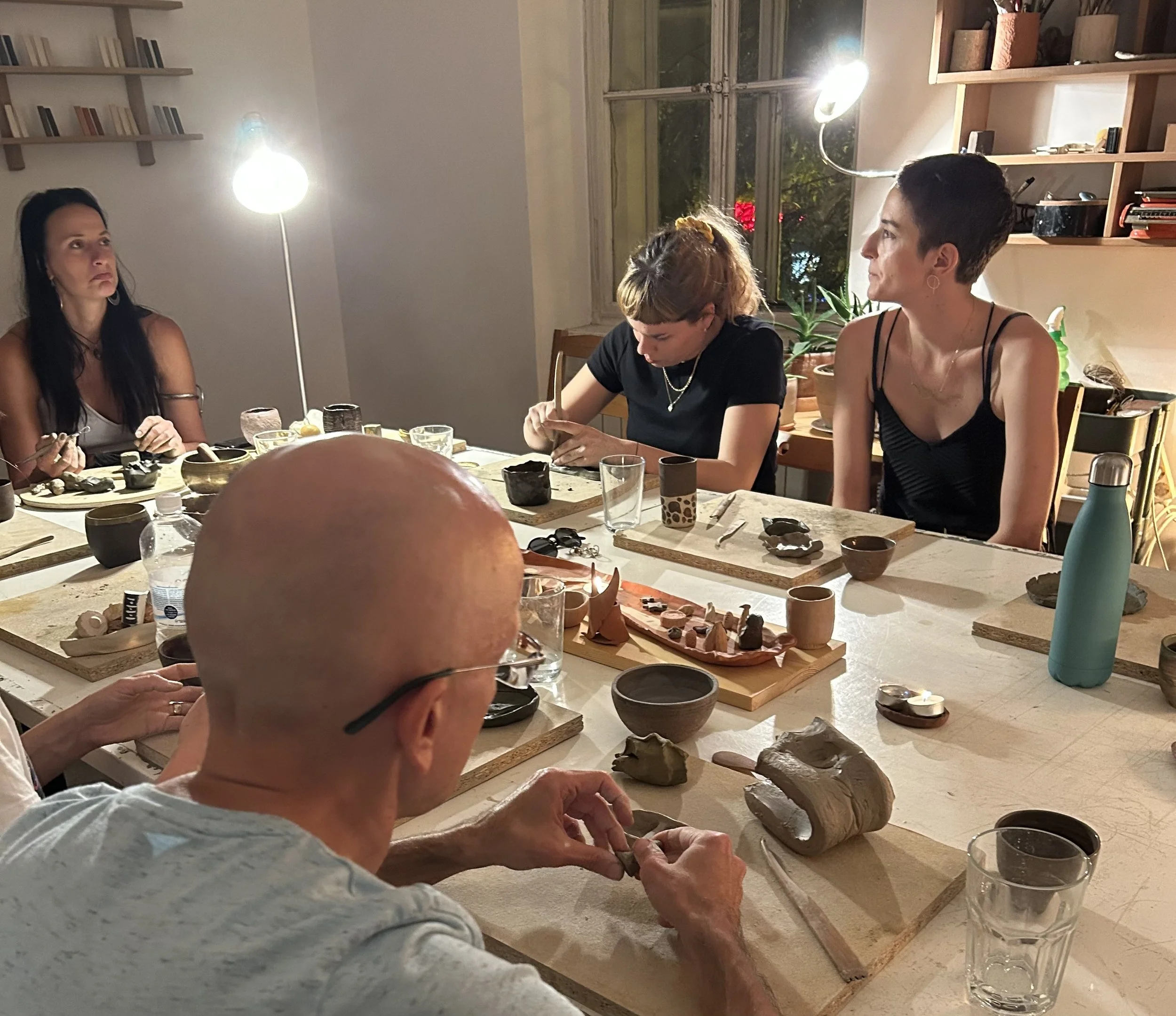 People sitting at a table with pottery and ceramics in a well-lit room, possibly engaging in pottery or ceramics workshop.