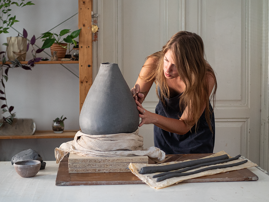 A woman working on a large, gray ceramic vase in a pottery studio, with pottery tools and supplies on the table.