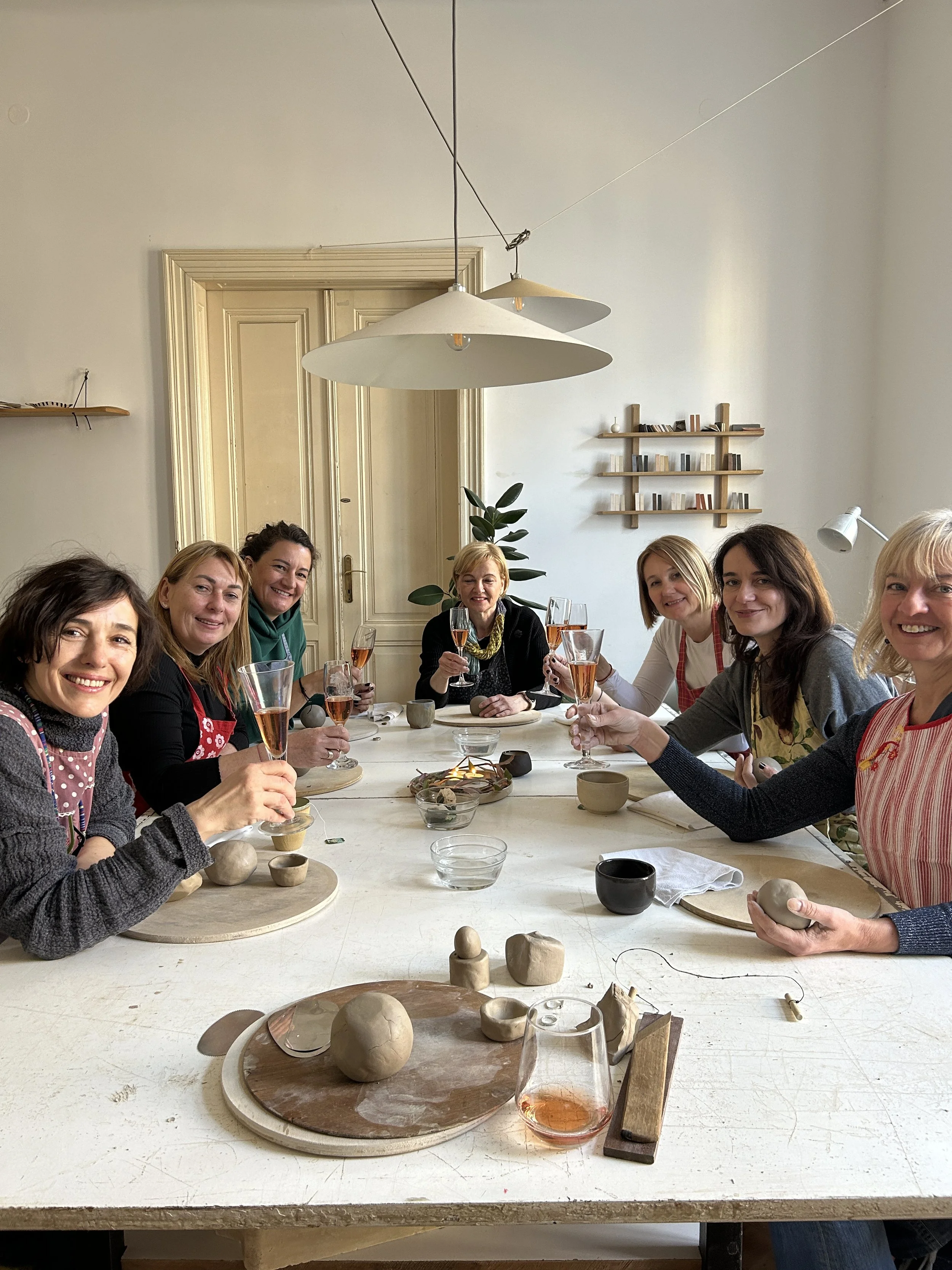 A group of women sitting around a table in a bright room, holding glasses of rosé wine, with art supplies and clay sculptures on the table, and a wooden shelf with books on the wall behind them.