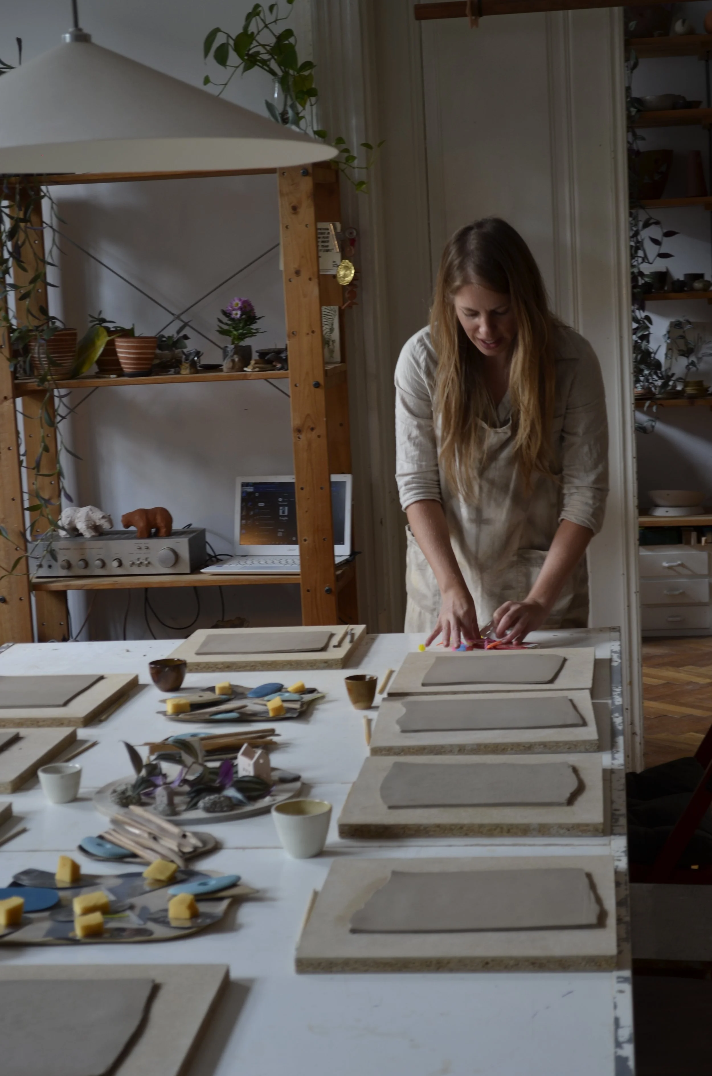 A woman working on a craft project at a table with various art supplies, including framed artwork, bowls, and tools. The workspace has wooden shelves with plants, bowls, and other decorative items.