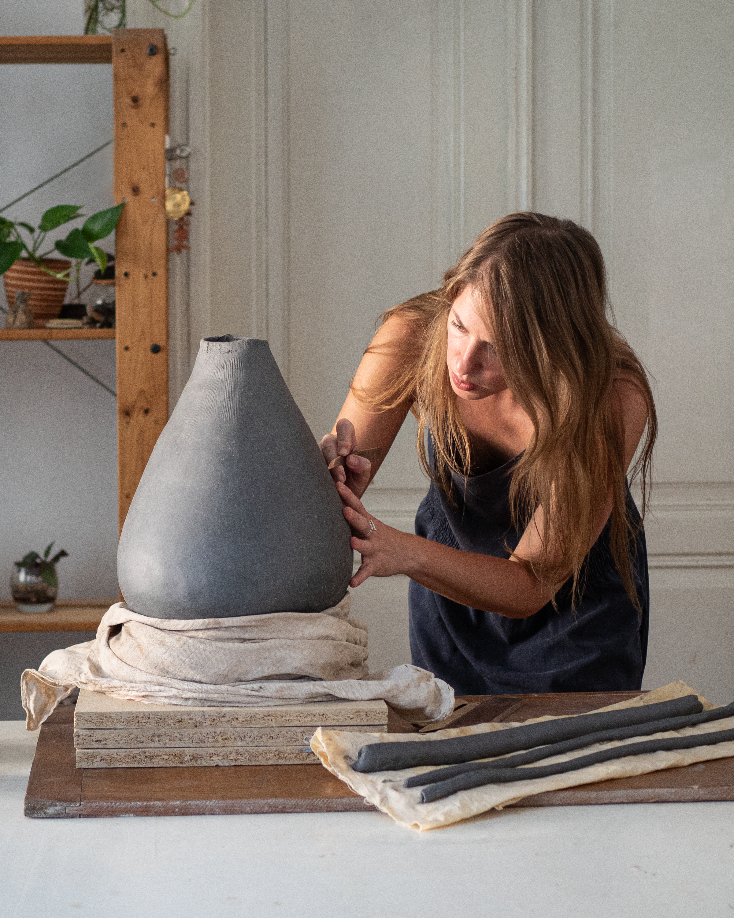 A woman is sculpting a large, rounded ceramic vase with a hand tool while leaning over a worktable covered with a cloth. The vase is placed on a layered wooden base. The workspace includes a wooden shelf with potted plants and small objects in the background.