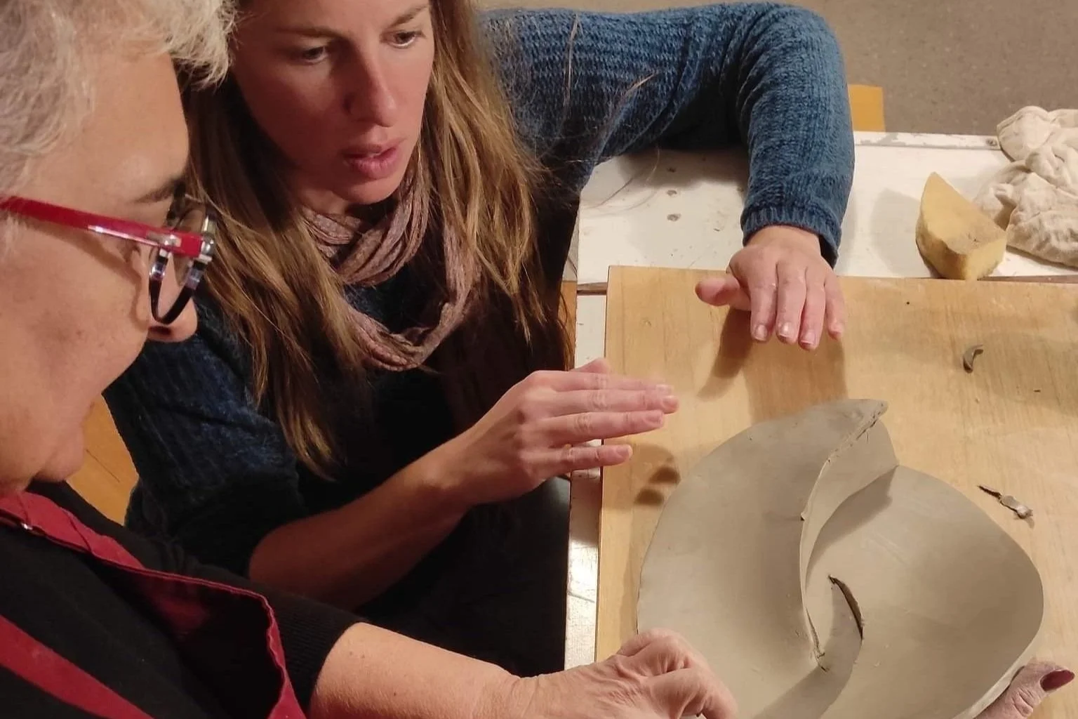 Two women examine a ceramic art piece on a wooden table, with one woman pointing and the other observing closely.