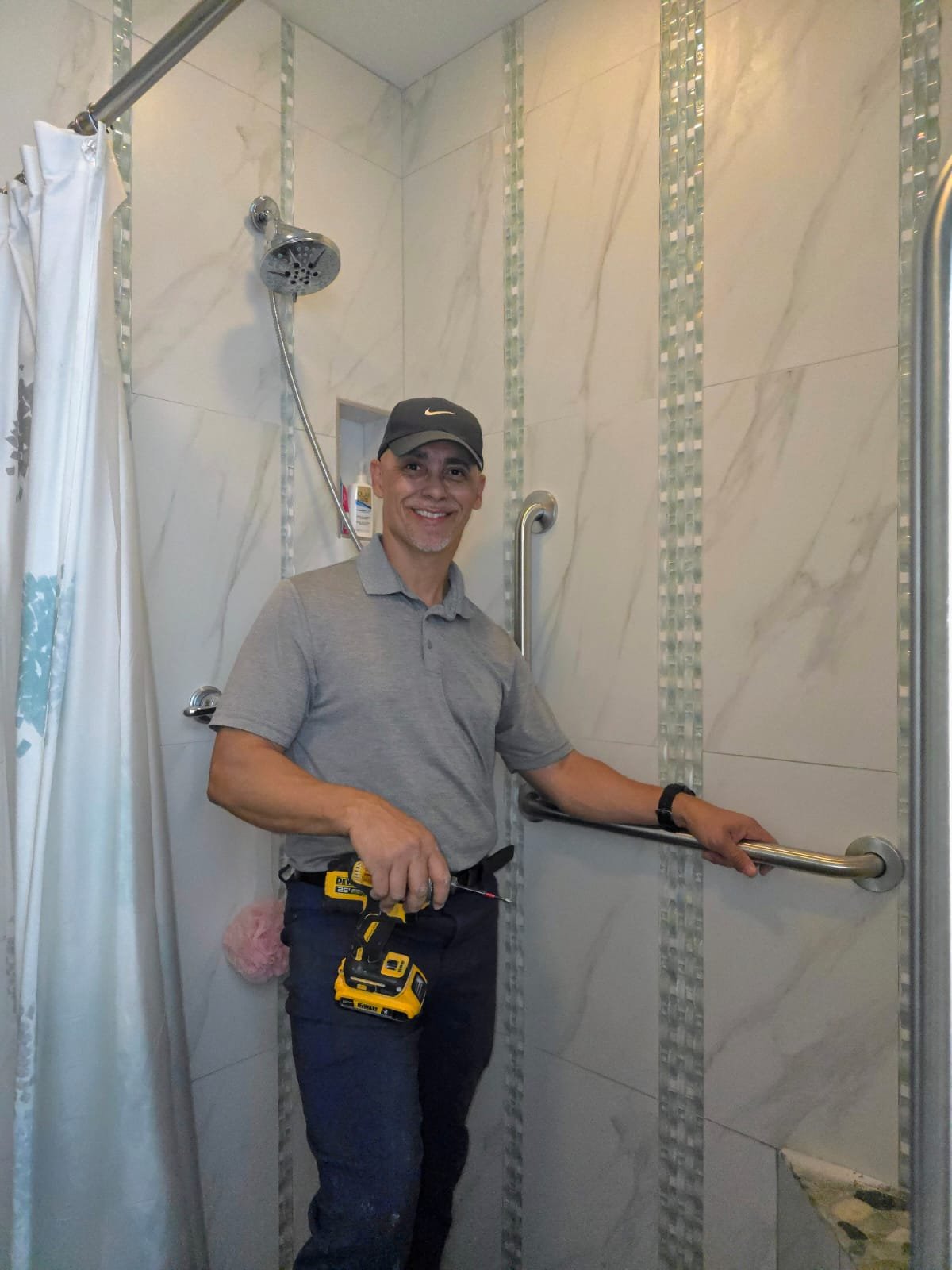 A man wearing a gray polo shirt, dark pants, and a black cap standing in a shower with marble walls, holding a cordless drill and smiling. He is installing or repairing safety grab bars in the shower.