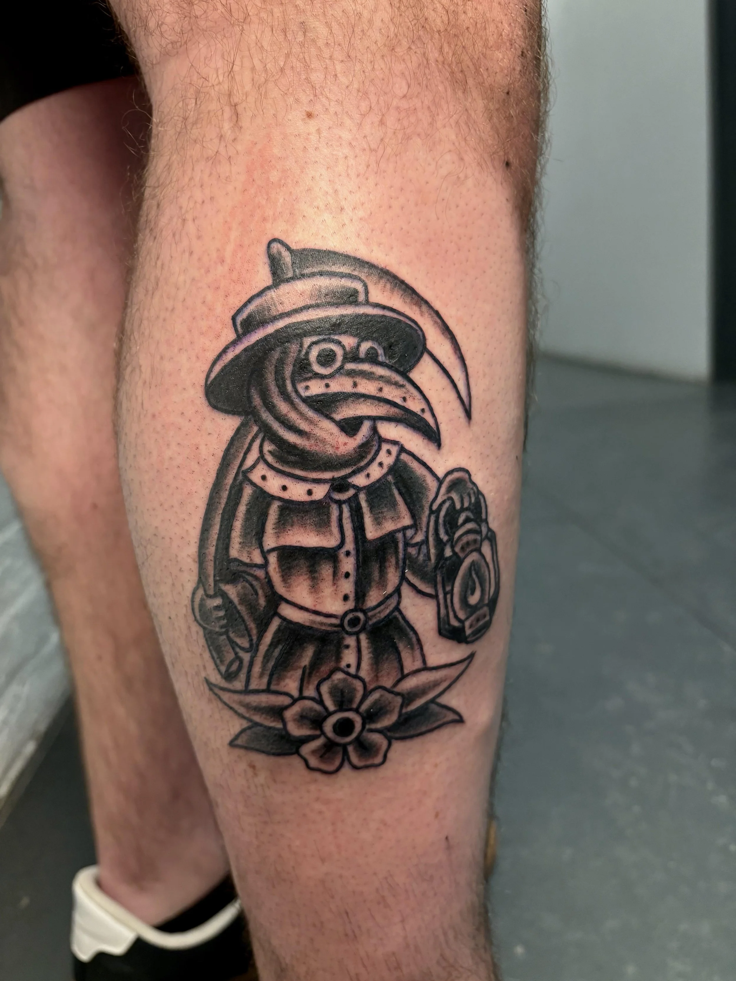 A black and gray plague doctor tattoo finished in an american traditional style. He is holding a lantern out and has a sythe leaning on his shoulder