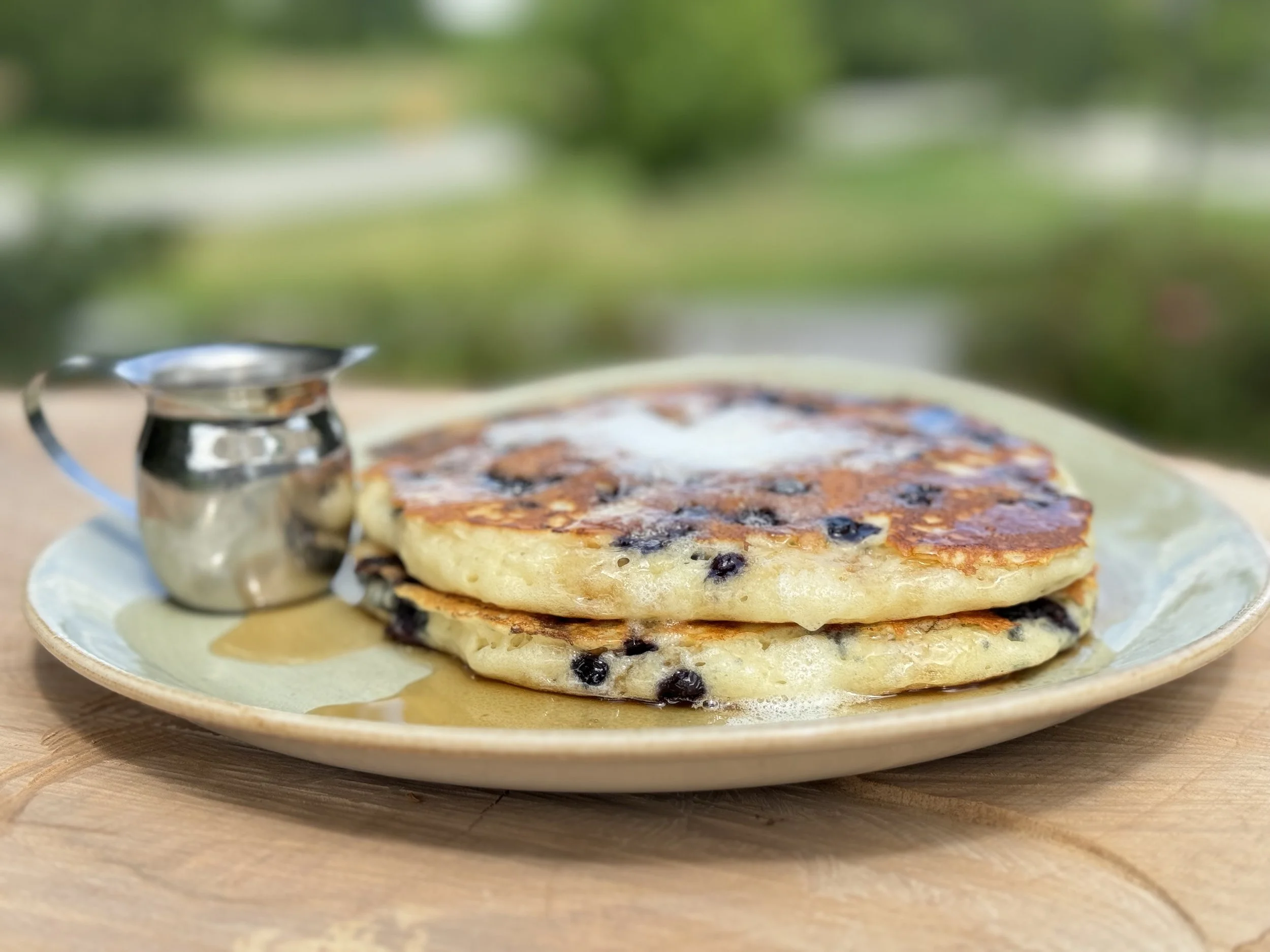 A stack of blueberry pancakes on a plate with a small silver jug of syrup, set on a wooden surface outdoors.