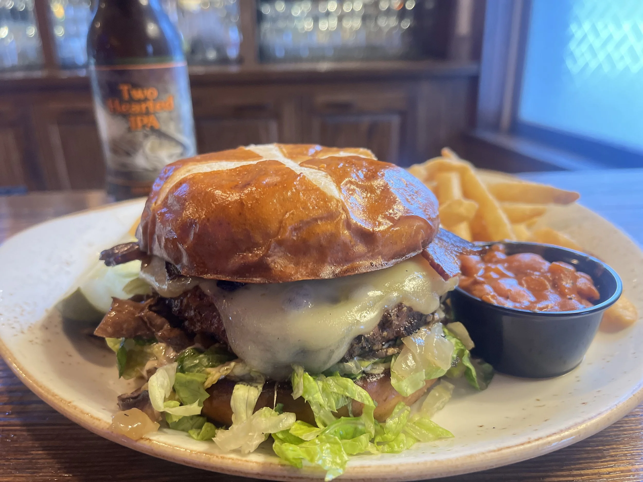 A cheeseburger on a pretzel bun with lettuce, served on a plate with a side of fries and a small cup of baked beans. A bottle of beer is in the background.
