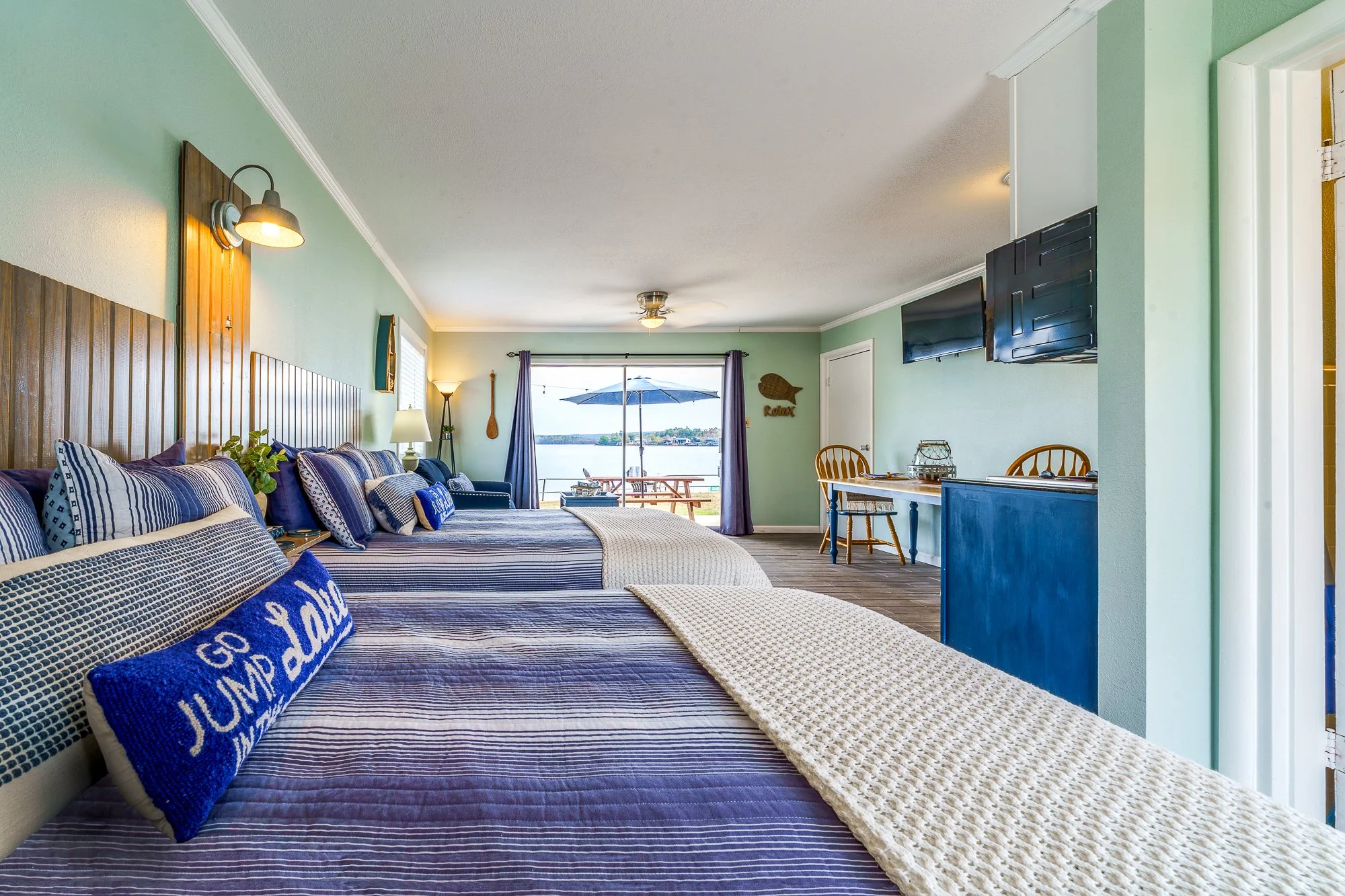 Nautical-themed hotel room with two beds, striped bedding, decorative pillows, a wall-mounted lamp, a TV, a small table with chairs, and a view of a lake through sliding glass doors with a patio outside.