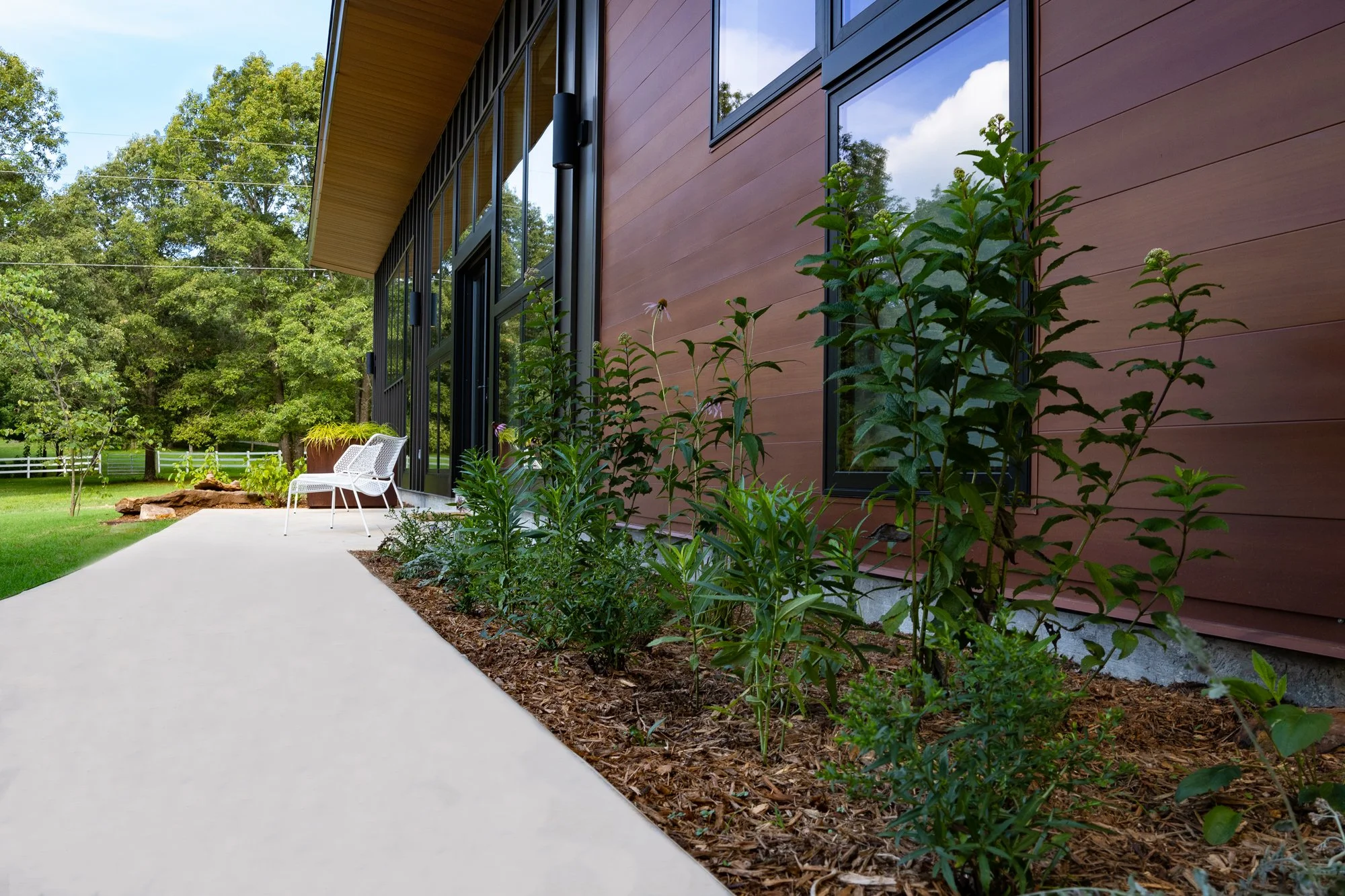 Exterior of modern house with wood siding, large windows, a garden with plants along the side, patio with chairs, and view of trees and lawn in the background.