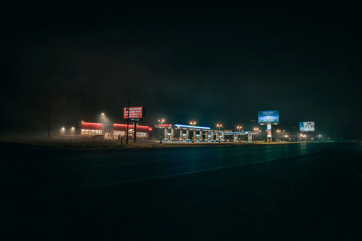 Nighttime scene of a gas station and convenience store illuminated by streetlights, with billboards in the background.