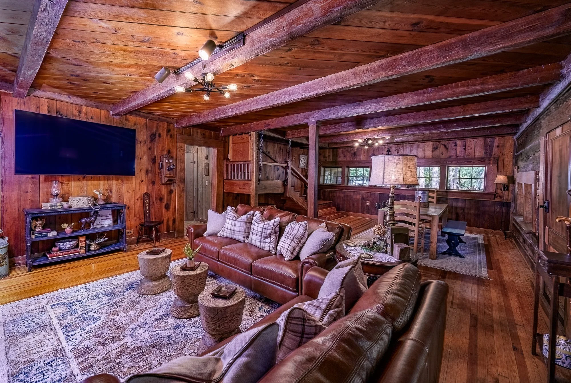 Rustic living room with leather sofas, wooden ceiling beams, and a large flat-screen TV. The space features wooden walls, a patterned area rug, small round tables, and decorative lighting. A dining area with a wooden table and chairs is visible in th