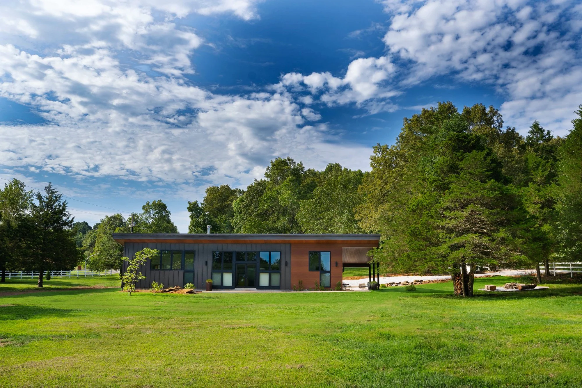 Modern ranch-style house in a lush green landscape with trees and a blue sky with clouds.