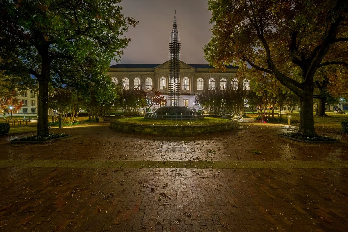 Night view of a grand building with arched windows, surrounded by trees and a brick pathway, with a tall, illuminated sculpture in the foreground.