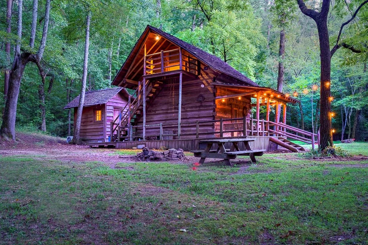 Rustic wooden cabin with porch and string lights, set in a forest with a picnic table and fire pit in the foreground.