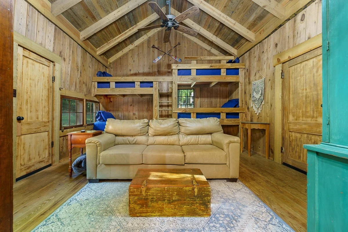 Cozy rustic cabin interior with wooden walls and ceiling, a leather couch, blue bunk beds, vintage trunk coffee table, and ceiling fan. Decor includes a rug, wall tapestry, and small wooden furniture pieces.