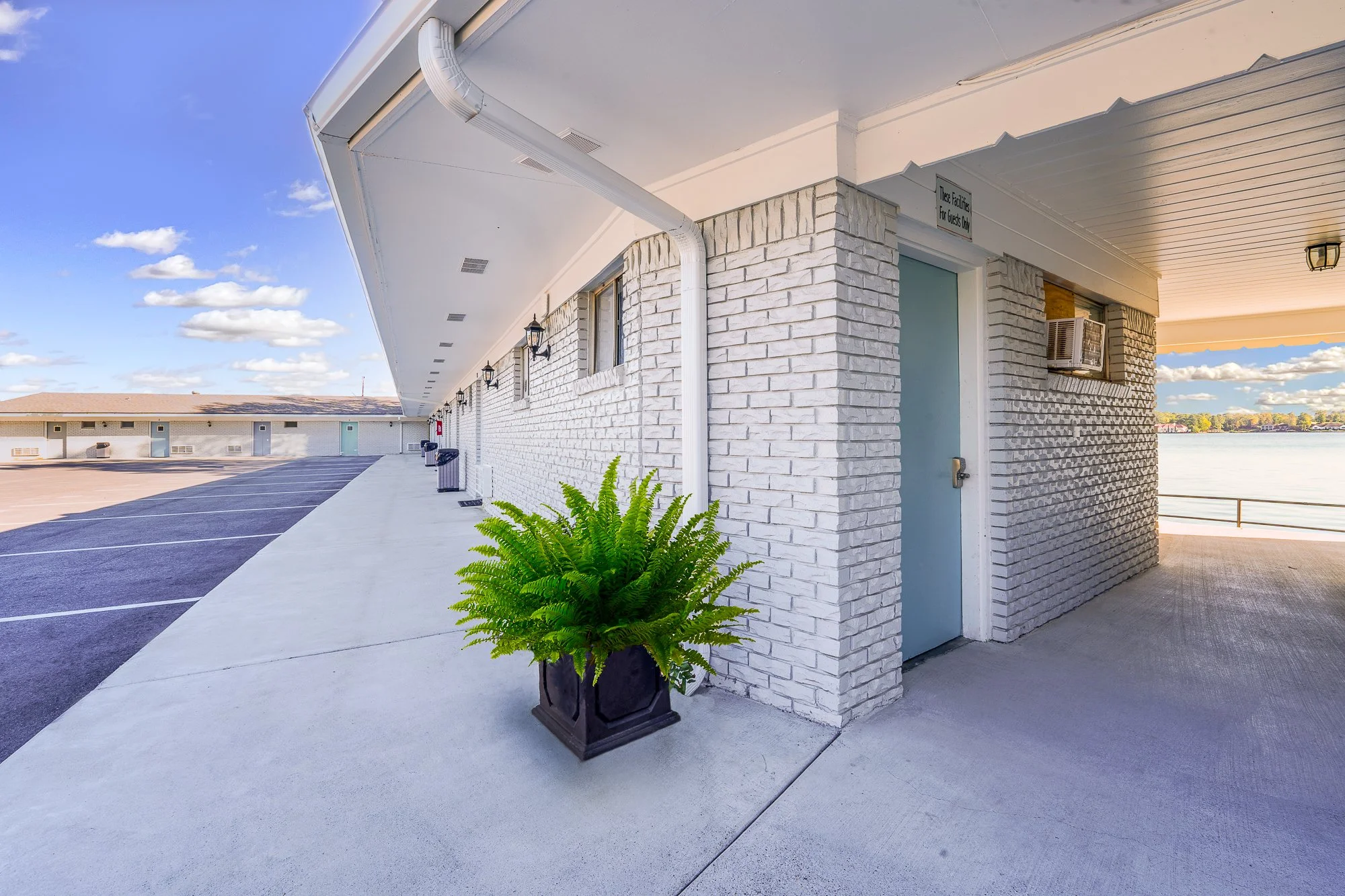 Exterior view of a motel with white brick walls, a blue door, a potted fern, and a view of a lake in the background. The motel has a long corridor with parking spaces and outdoor lighting.