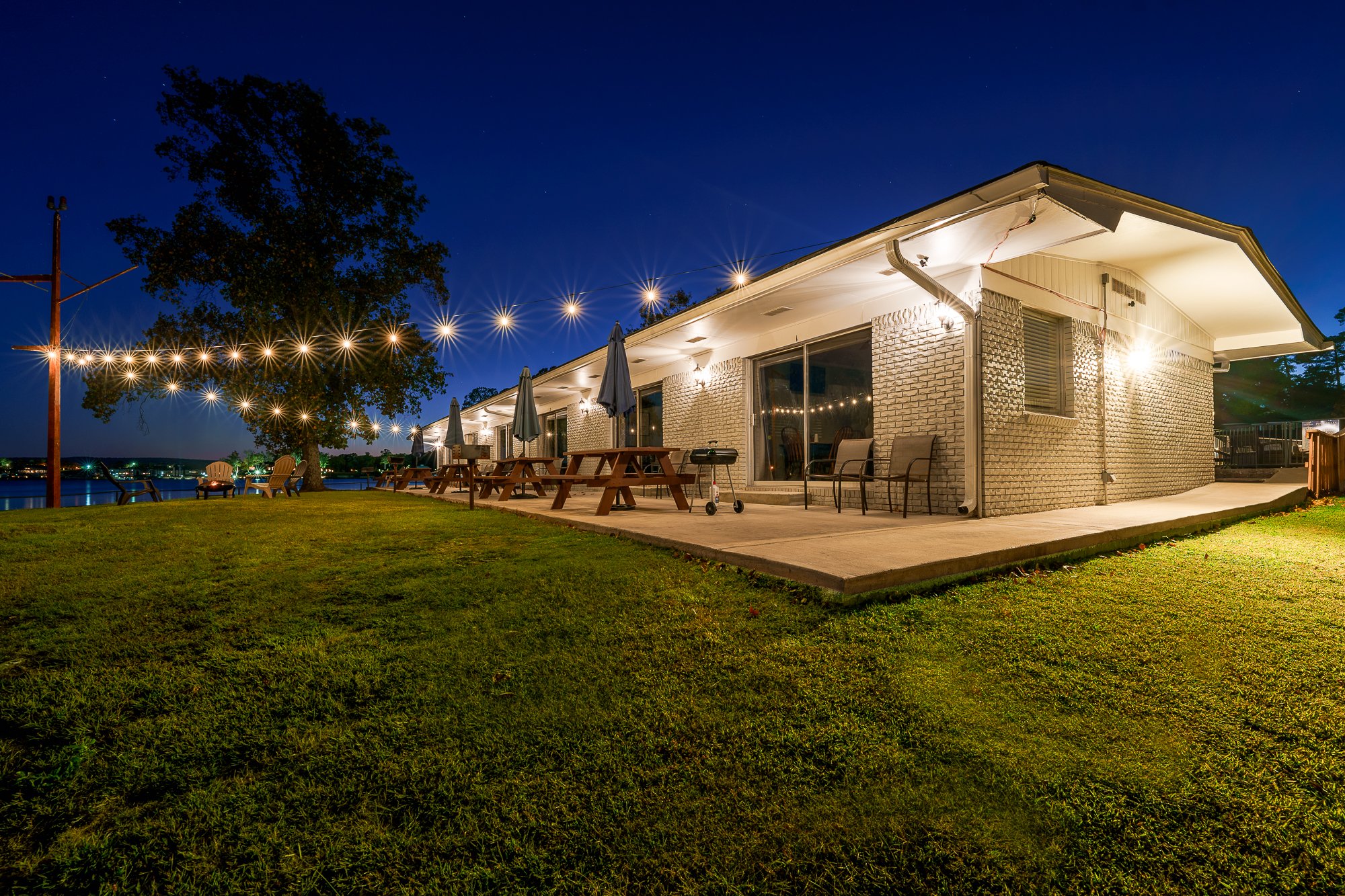 Exterior view of a house with string lights at night, featuring a well-lit patio with picnic tables and chairs, adjacent to a green lawn and a tree, overlooking water.
