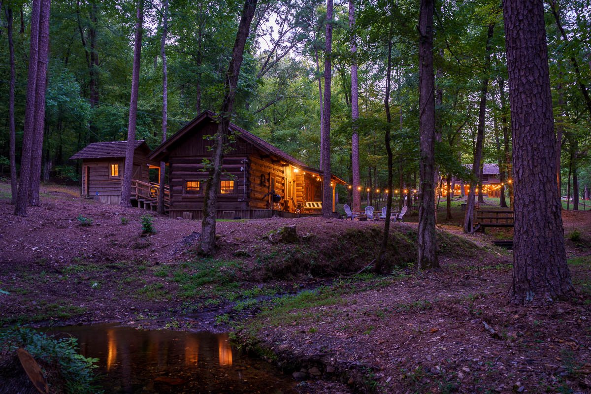 Rustic log cabin in a wooded area at dusk, illuminated by warm lights and surrounded by trees.
