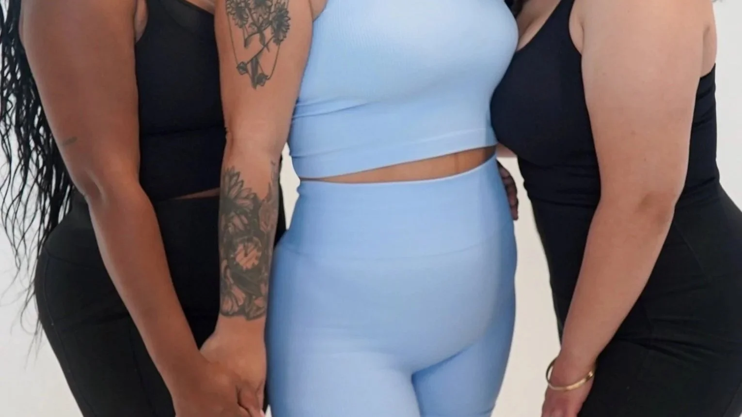 Close-up of three women standing together, wearing sleeveless tops and form-fitting pants.