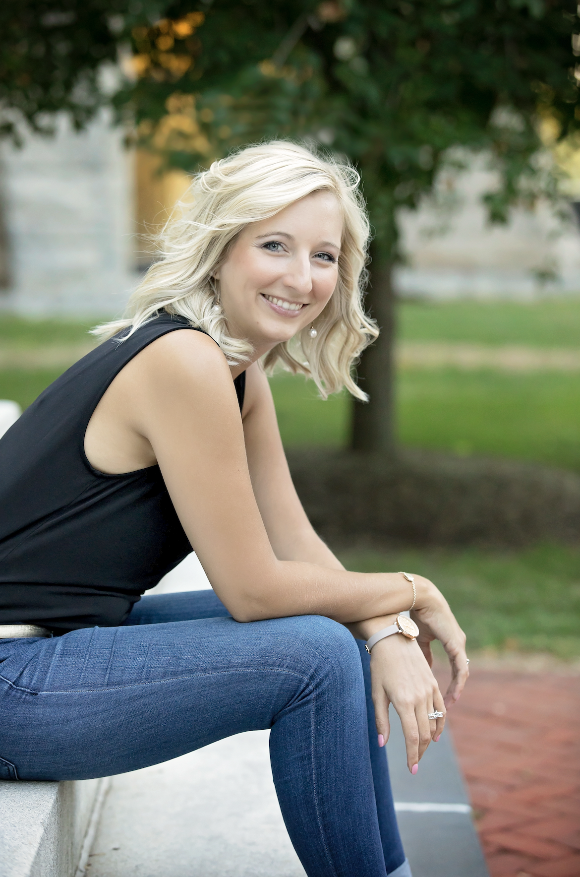 Smiling person with blonde hair sitting outdoors on a bench, wearing a black sleeveless top and jeans, with trees in the background.