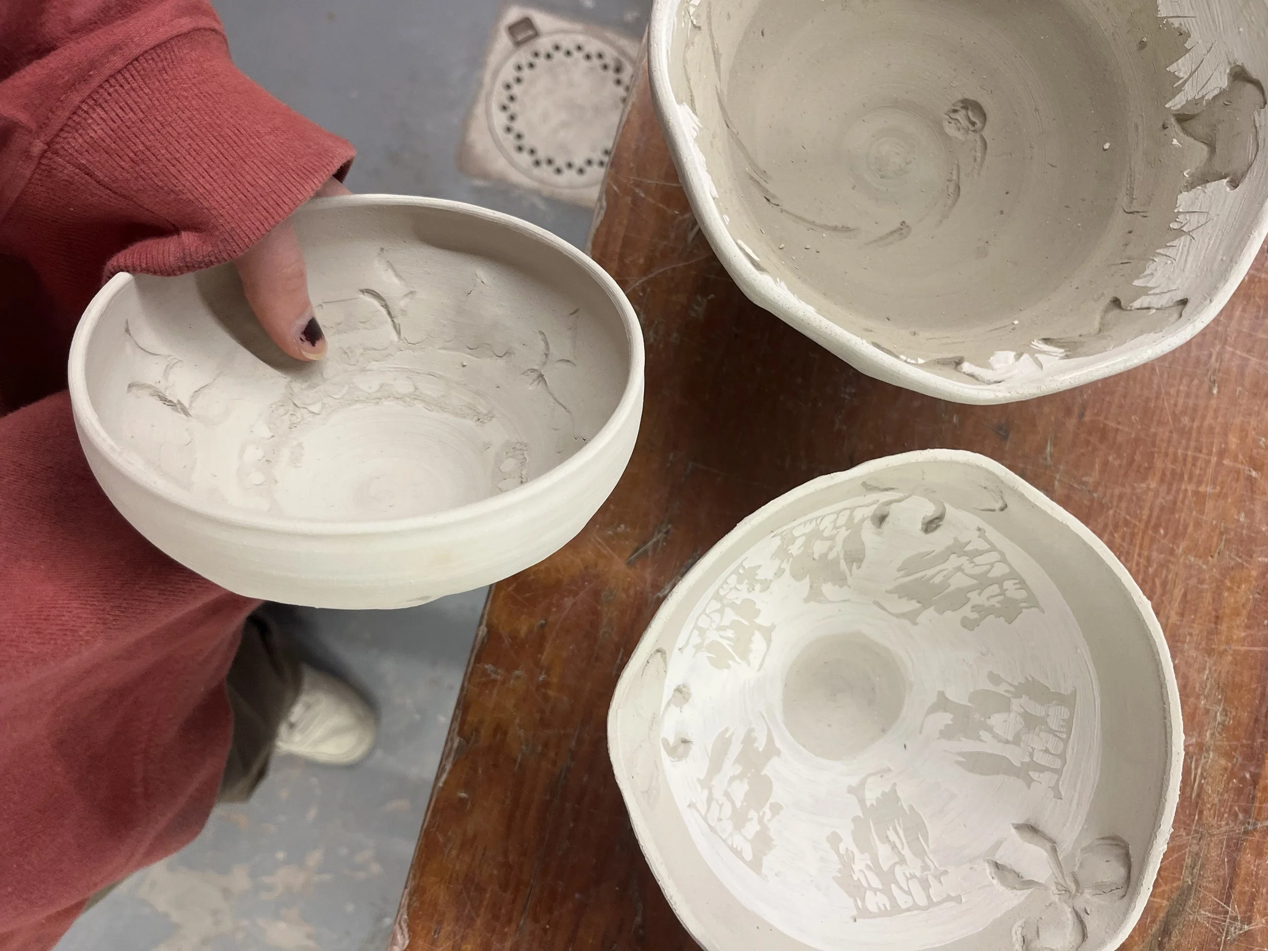 Three unfinished ceramic bowls, two placed on a wooden work surface and one being held by a person wearing a red long-sleeve shirt. The bowls are hollow with rough, unglazed interiors.