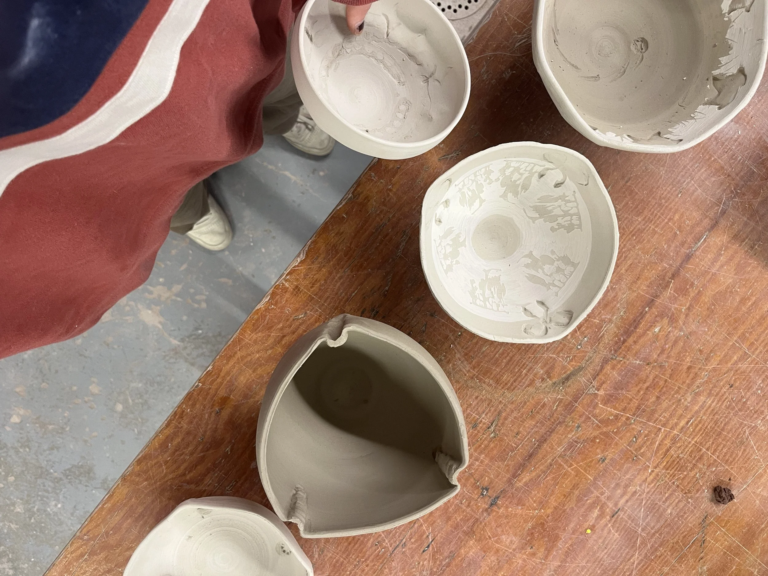 Several unglazed ceramic bowls on a wooden table, with some broken pieces and a person in a red and blue jacket next to them.