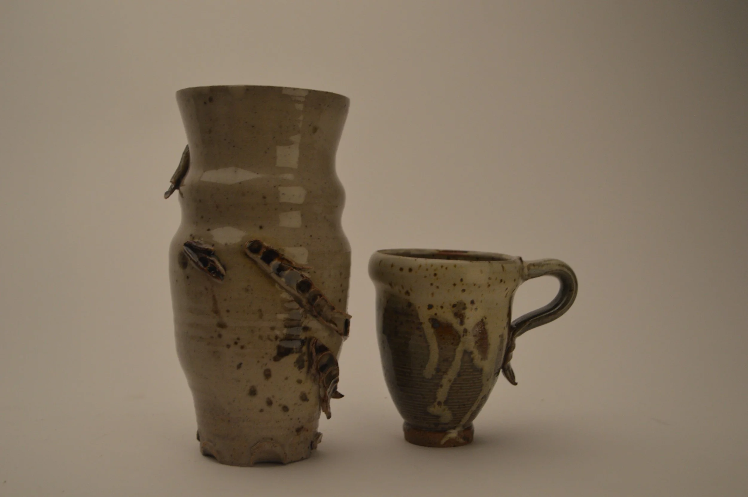 Two handmade ceramic vases with a speckled beige glaze, one tall and wavy with attached small glass-like sculptures, and the other short with a looped handle, against a plain background.