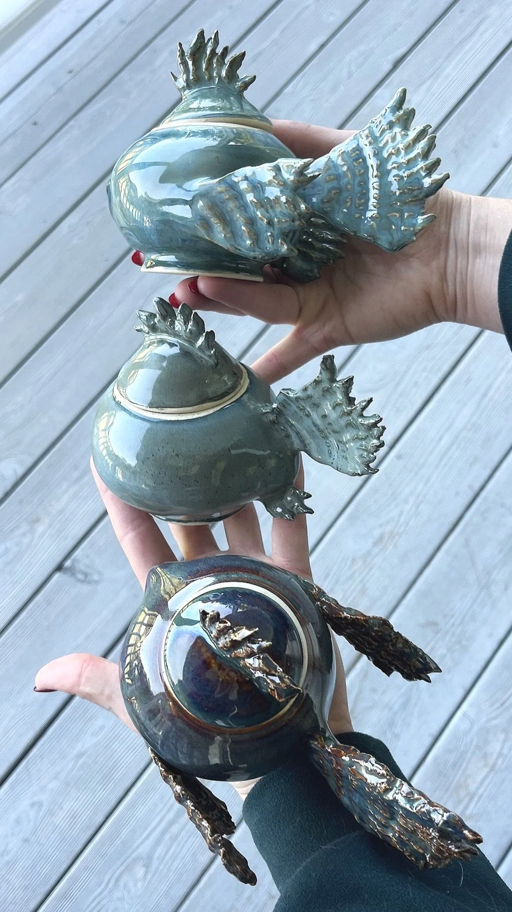 Three ceramic fish-shaped sculptures with detailed fins, held by hands over wooden planks.