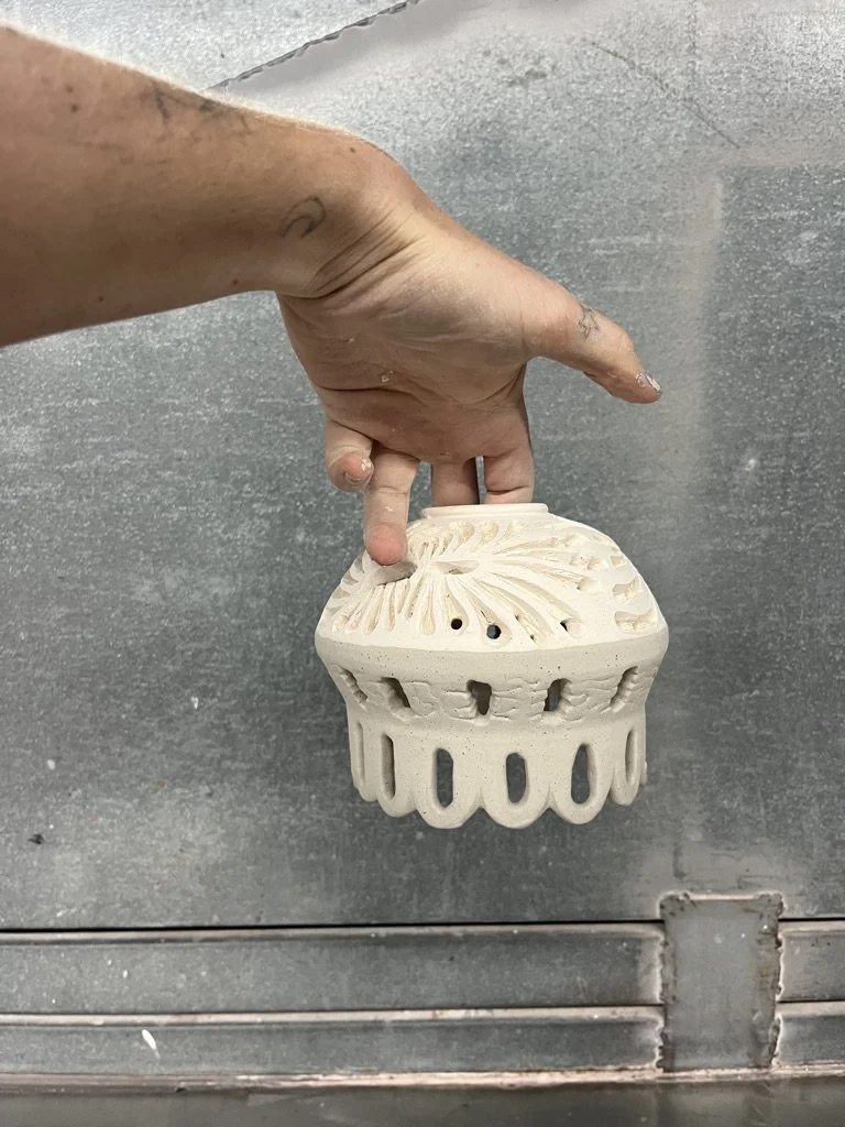 A hand holding a partially finished ceramic piece with intricate carved details and holes, likely part of a pottery or sculpture project.