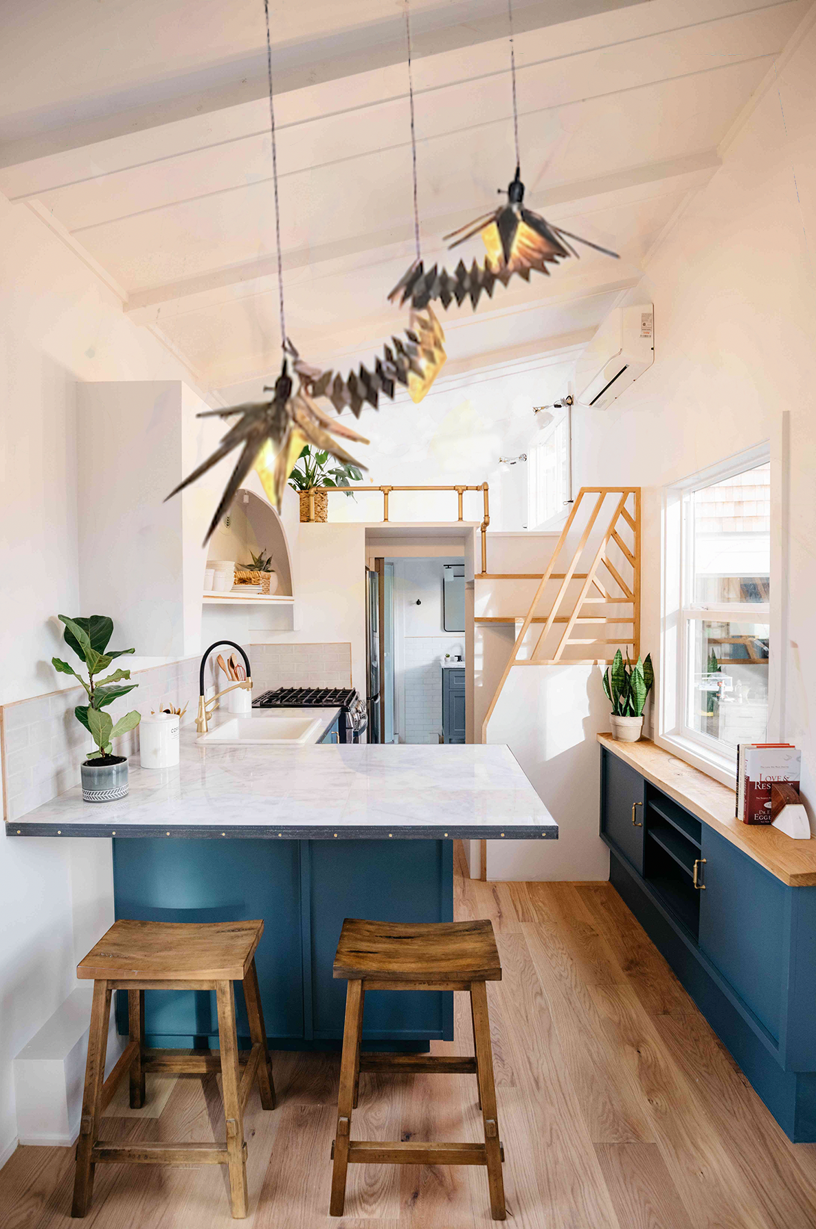 Interior view of a modern kitchen with blue lower cabinets, two wooden bar stools, a white marble countertop, and potted plants. Wooden stairs lead up to a loft area with a wooden railing. A decorative bird-shaped hanging mobile is suspended from the ceiling.
