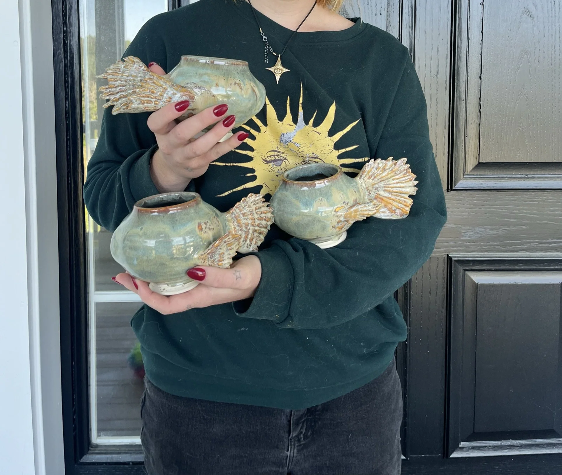 Person holding three fish-shaped ceramic pots with open tops, dark green sweatshirt with a sun graphic, black jeans, and red nail polish, standing in front of a black door.
