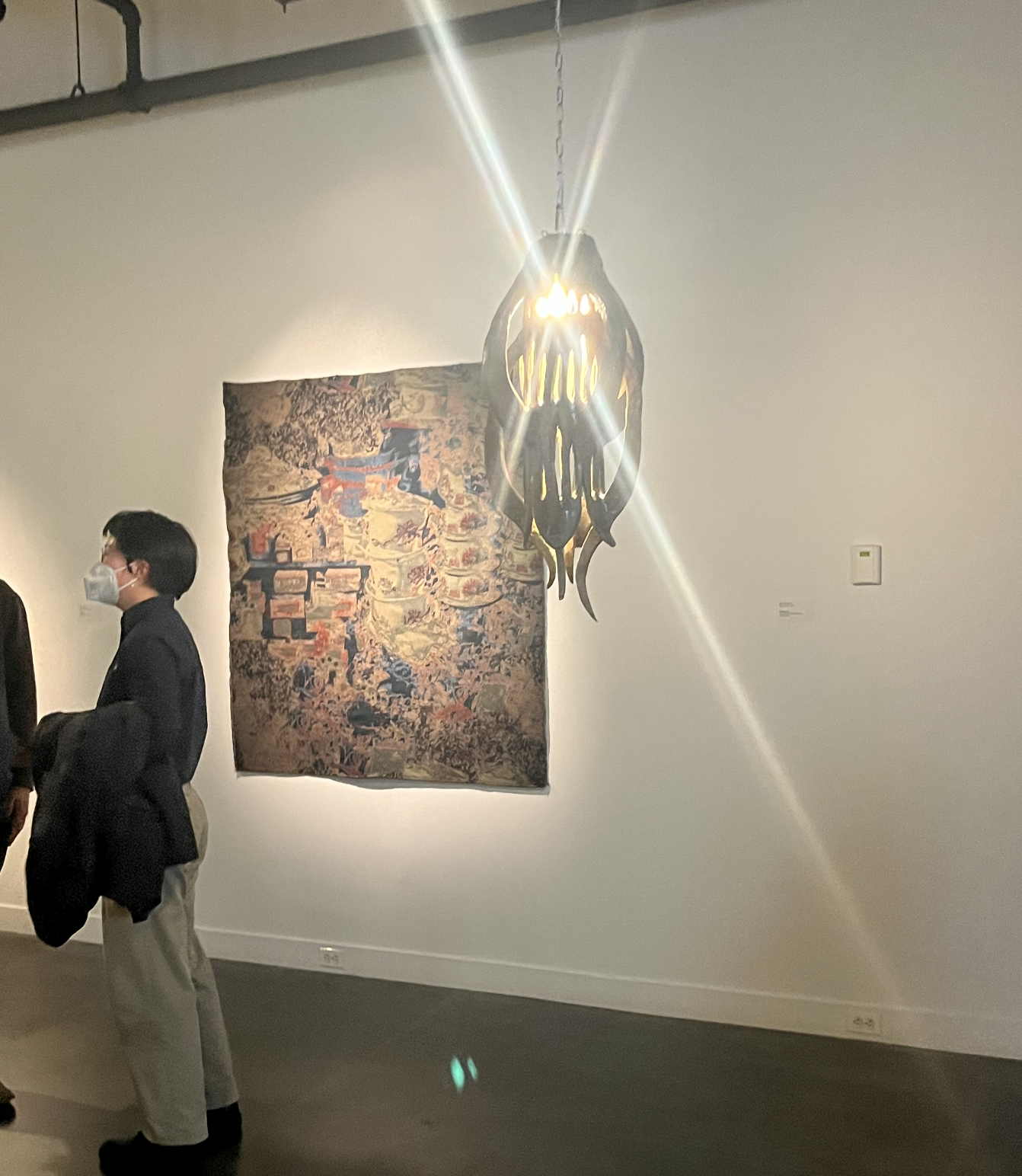 People wearing masks in an art gallery, looking at a colorful abstract painting and a hanging sculpture with light shining through.