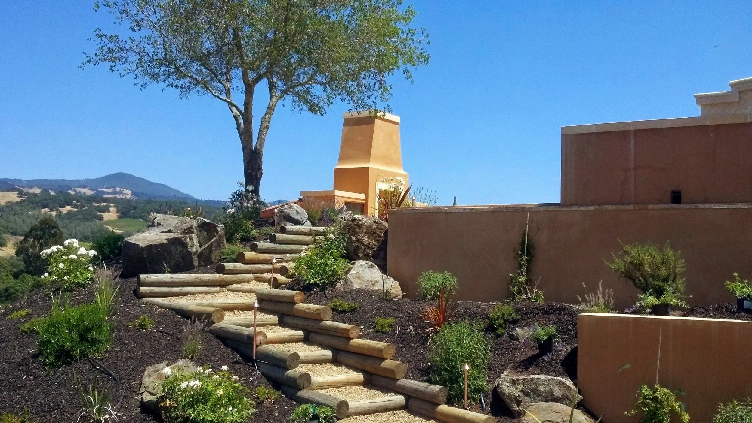 Manicured garden with wooden log and sand steps leading to wood burning fireplace on a pavered patio