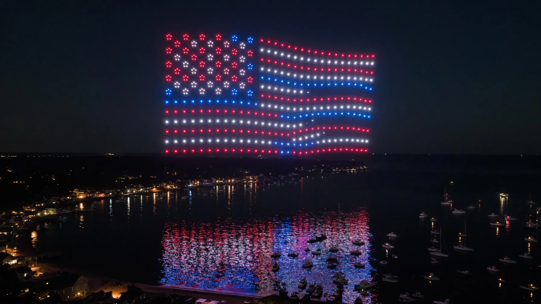 Hundreds of synchronized drones forming a glowing American flag in red, white, and blue over a harbor at night, with the formation reflected on the water below