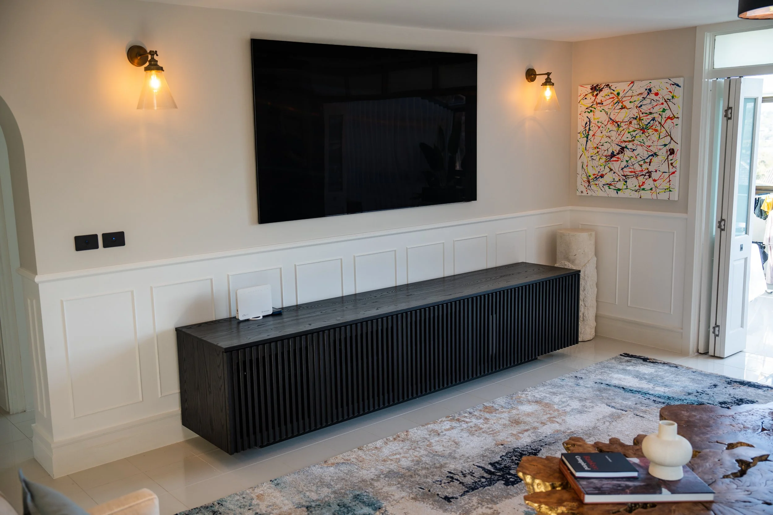 Living room with white walls, wall-mounted TV, two industrial-style wall sconces, abstract colorful painting, black wooden TV stand, area rug, coffee table with a decorative white vase and books, and an open door leading outside.