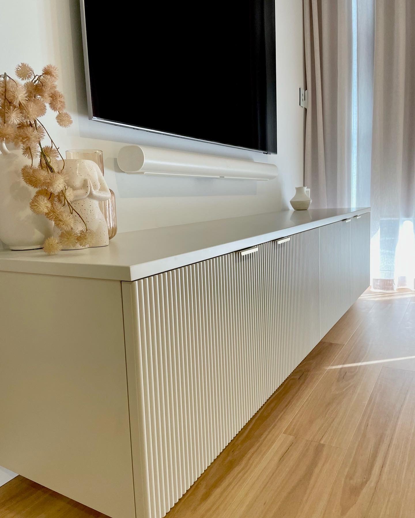 White modern TV stand with textured vertical ridges, decorated with white vases and dried flowers, in front of a wall-mounted flat-screen television.