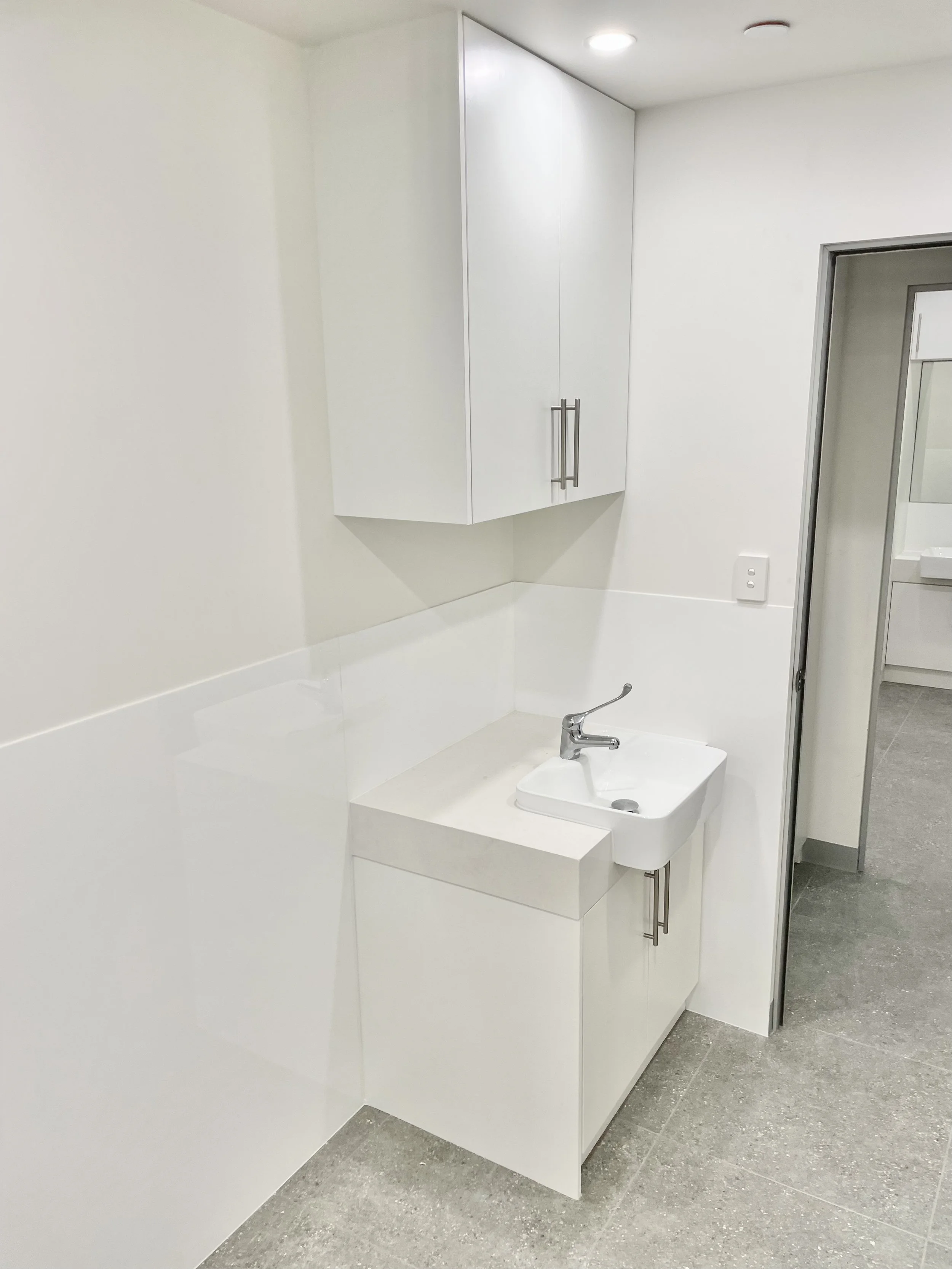 Small white utility sink with a faucet in a clean, minimalistic bathroom or laundry room with white walls and gray tile floor.