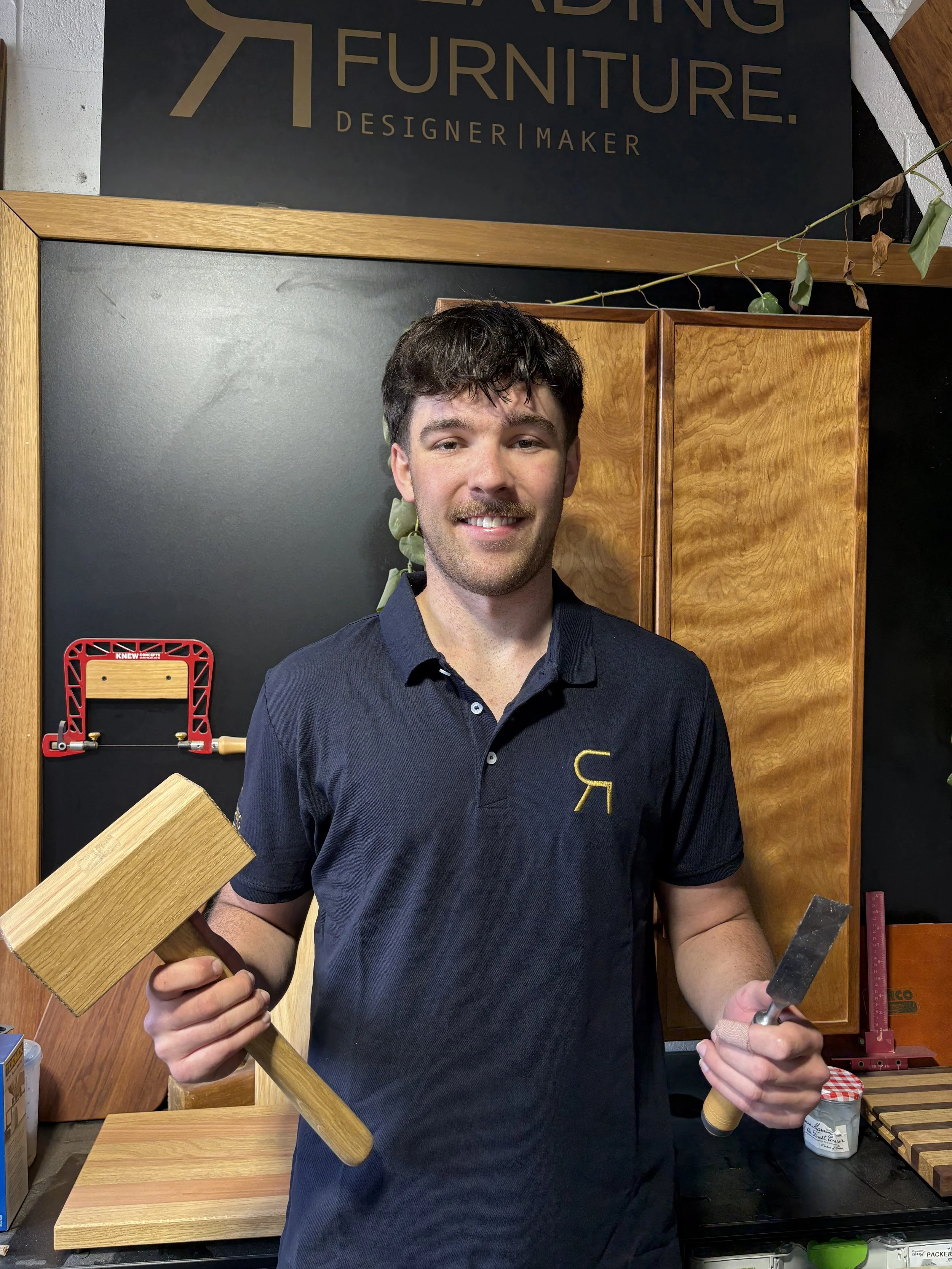A young man with dark hair and a beard smiling, holding a wooden mallet in his left hand and a chisel in his right hand, standing in a woodworking shop with wooden cabinets and a black wall with a logo that reads 'READING FURNITURE. DESIGNER | MAKER.'